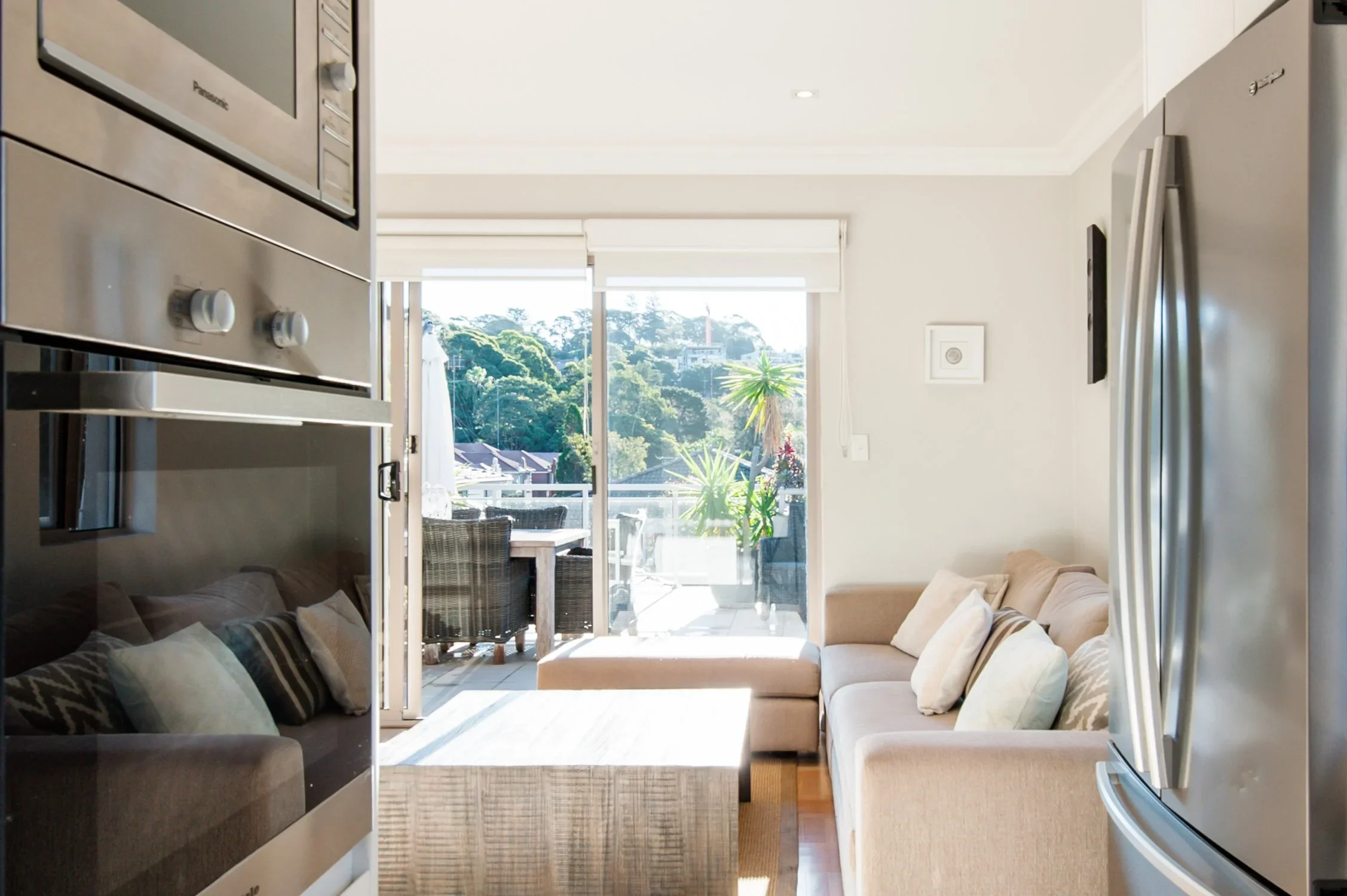 Living room with beige sectional sofa, coffee table, and sliding glass door leading to balcony with outdoor furniture and greenery.