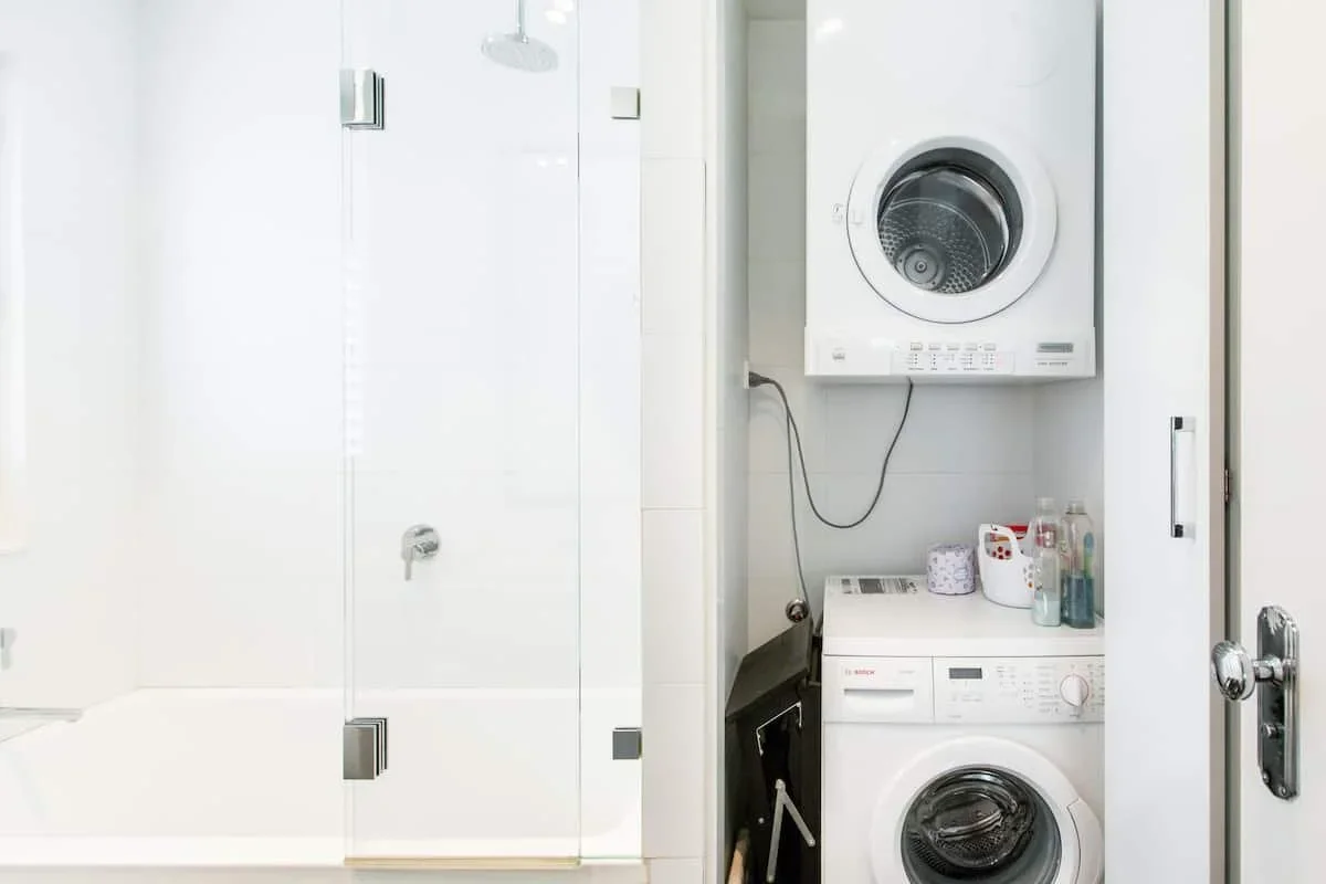 A small laundry area with a stacked washer and dryer, bottles of laundry detergent, a roll of toilet paper, a black bag, and a door leading to a shower or bathroom.