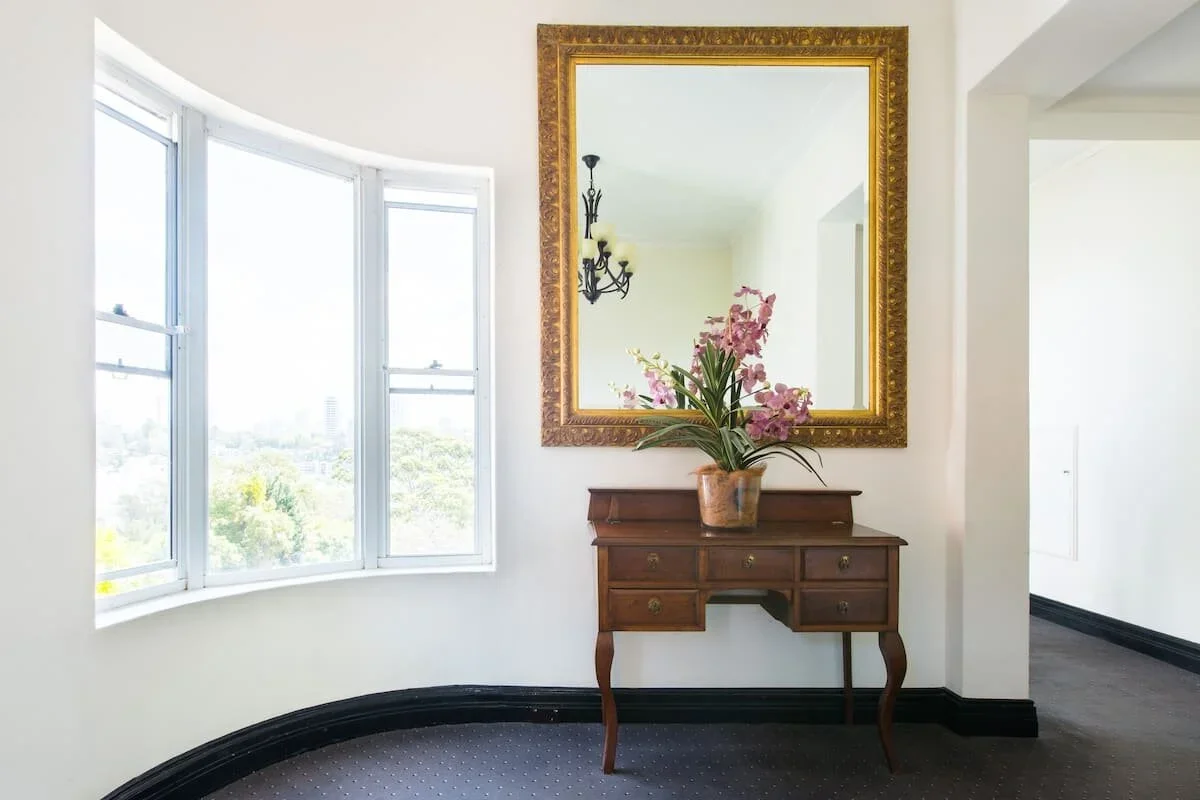An interior corner of a room with a window, a vintage wooden desk with four drawers, a potted pink orchid, a large gold-framed mirror, and a chandelier reflected in the mirror.