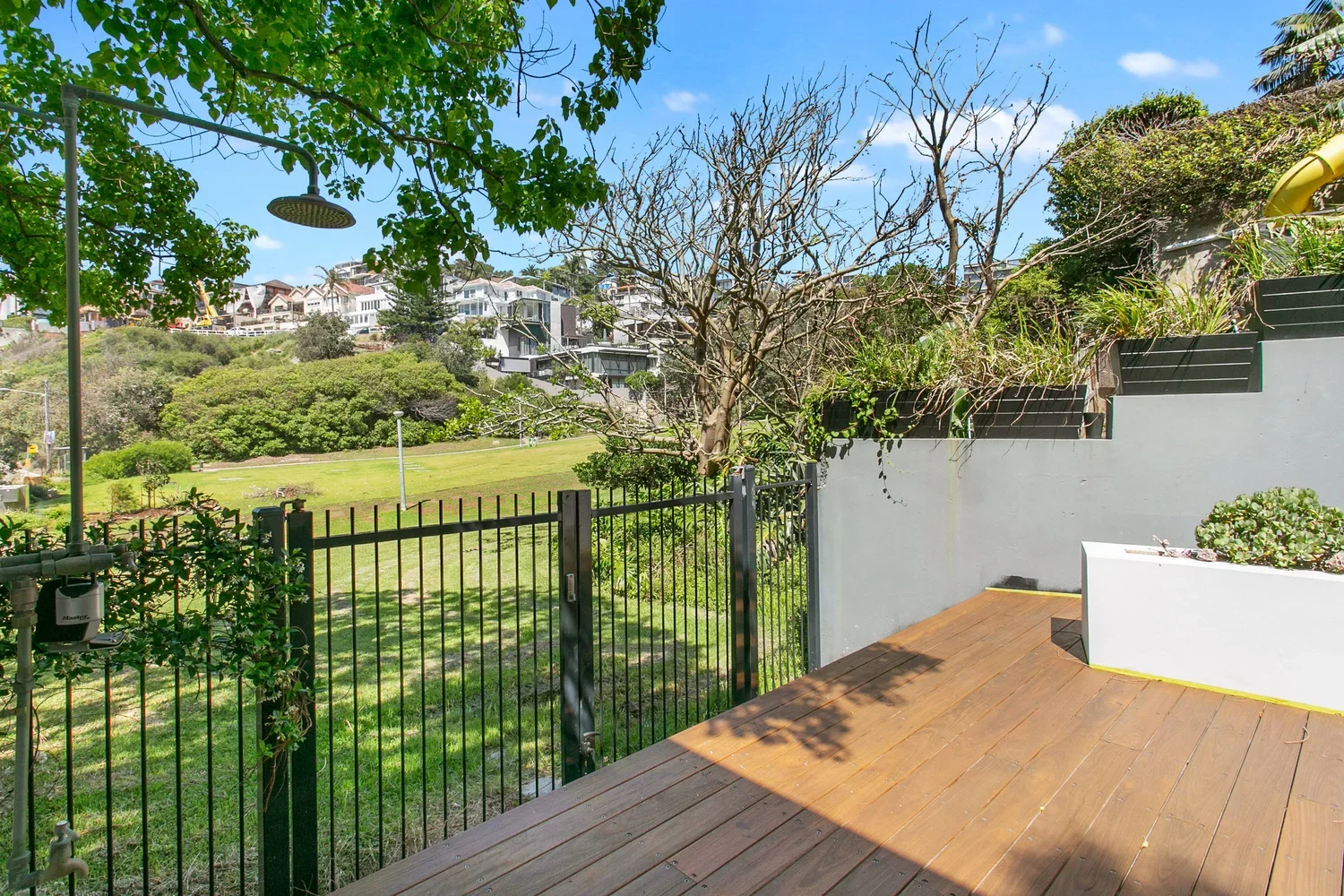 A backyard patio with wooden decking enclosed by a black metal fence, overlooking a green lawn with trees and residential houses in the distance under a blue sky with some clouds.