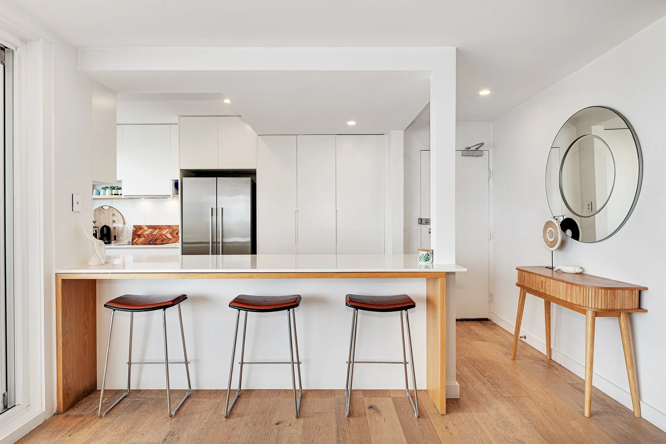 Modern kitchen with white walls, wooden accents, and a breakfast bar with three black and brown stools. There is a small wooden console table with decorative items and a large round mirror on the wall.