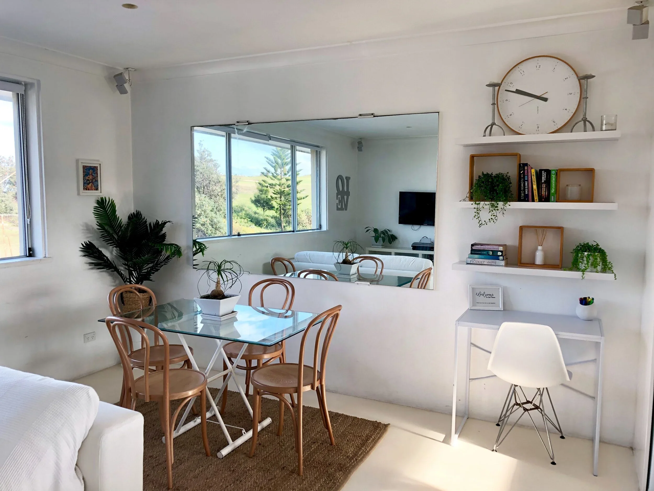 Bright dining room with glass-top table, six wooden chairs, potted plants, large wall mirror, and a white side desk with shelves holding books and plants. Large window with outdoor view, and a white sofa partially visible.