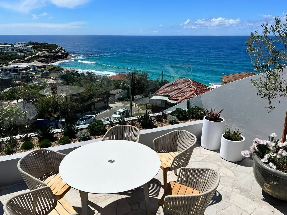 Outdoor patio with a round white table and six beige chairs overlooking a scenic ocean view. Potted plants and succulents decorate the patio, with a clear blue sky and the ocean in the background.