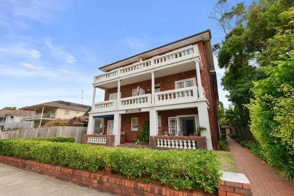 A three-story red brick building with white balcony railings and columns, surrounded by green bushes and trees, under a blue sky.