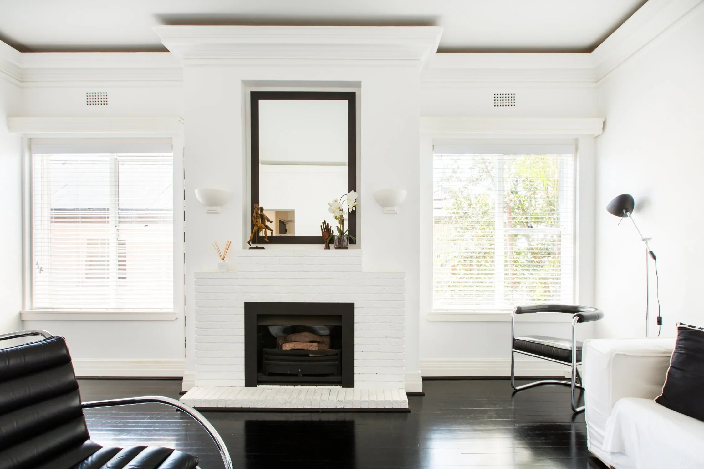 Bright living room with white walls, black floor, white brick fireplace, and large windows with blinds, furnished with black and white chairs and a white sofa, with a floor lamp.