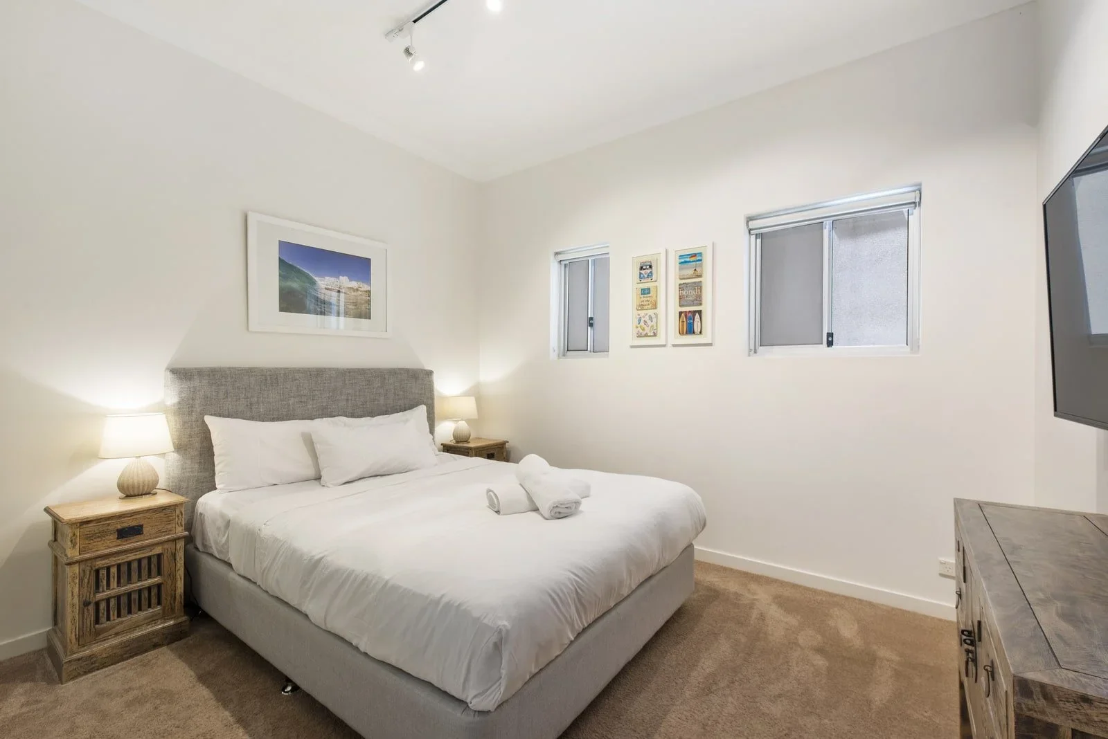 A neatly made bed in a minimalist hotel room with light-colored walls, two nightstands with lamps, framed artwork on the wall, and two small windows.