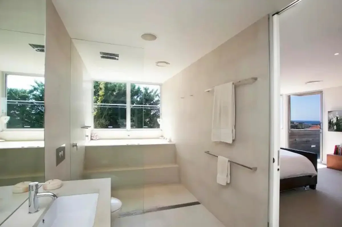 A modern bathroom with a large window, white walls, and minimalistic fixtures. There are two towels hanging on a rail, a sink, and a glass shower enclosure. Through the open door, a bedroom with a bed, window, and ocean view is visible.
