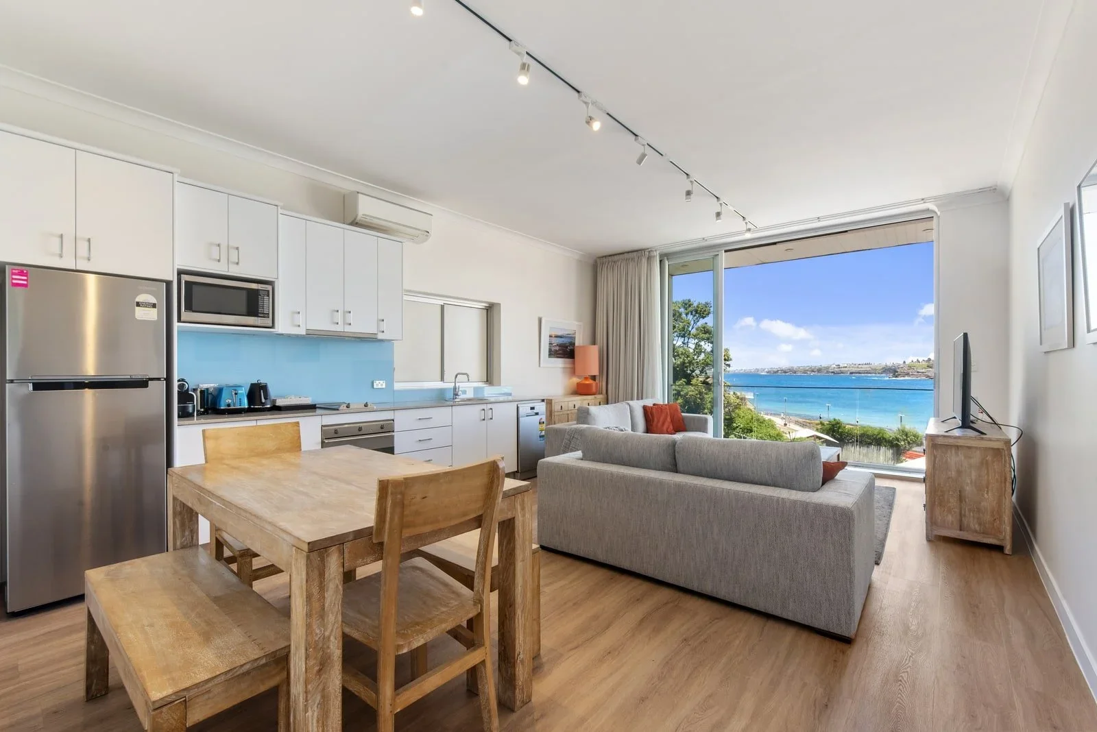 Open-concept living room and kitchen with large window overlooking a body of water, featuring white kitchen cabinets, stainless steel appliances, a small dining table with chairs, a gray sofa, and a TV on a wooden stand.
