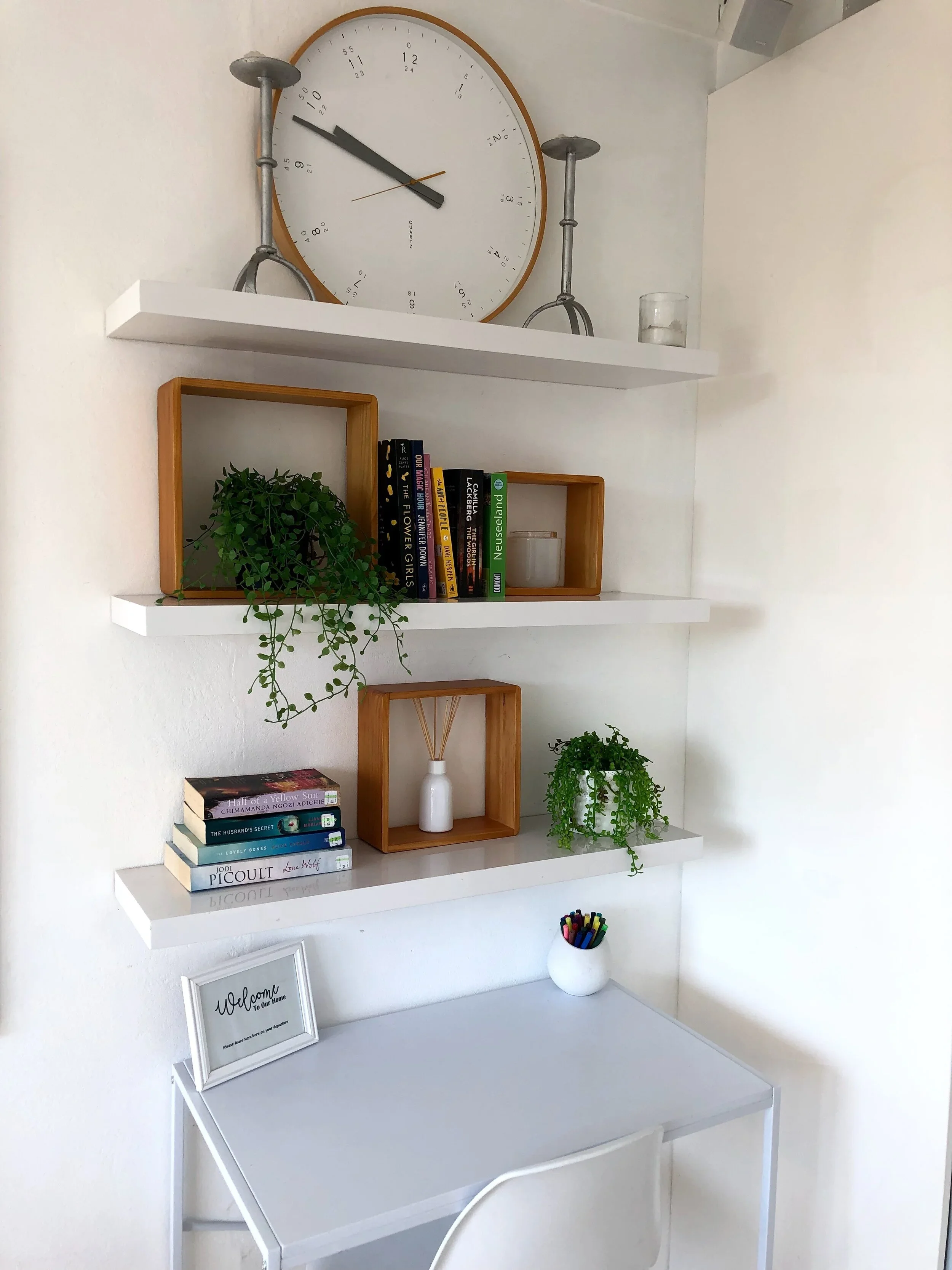 Wall-mounted shelves holding books, decorative plants, a framed sign, and a small container of markers, with a large wall clock above and a white desk below.