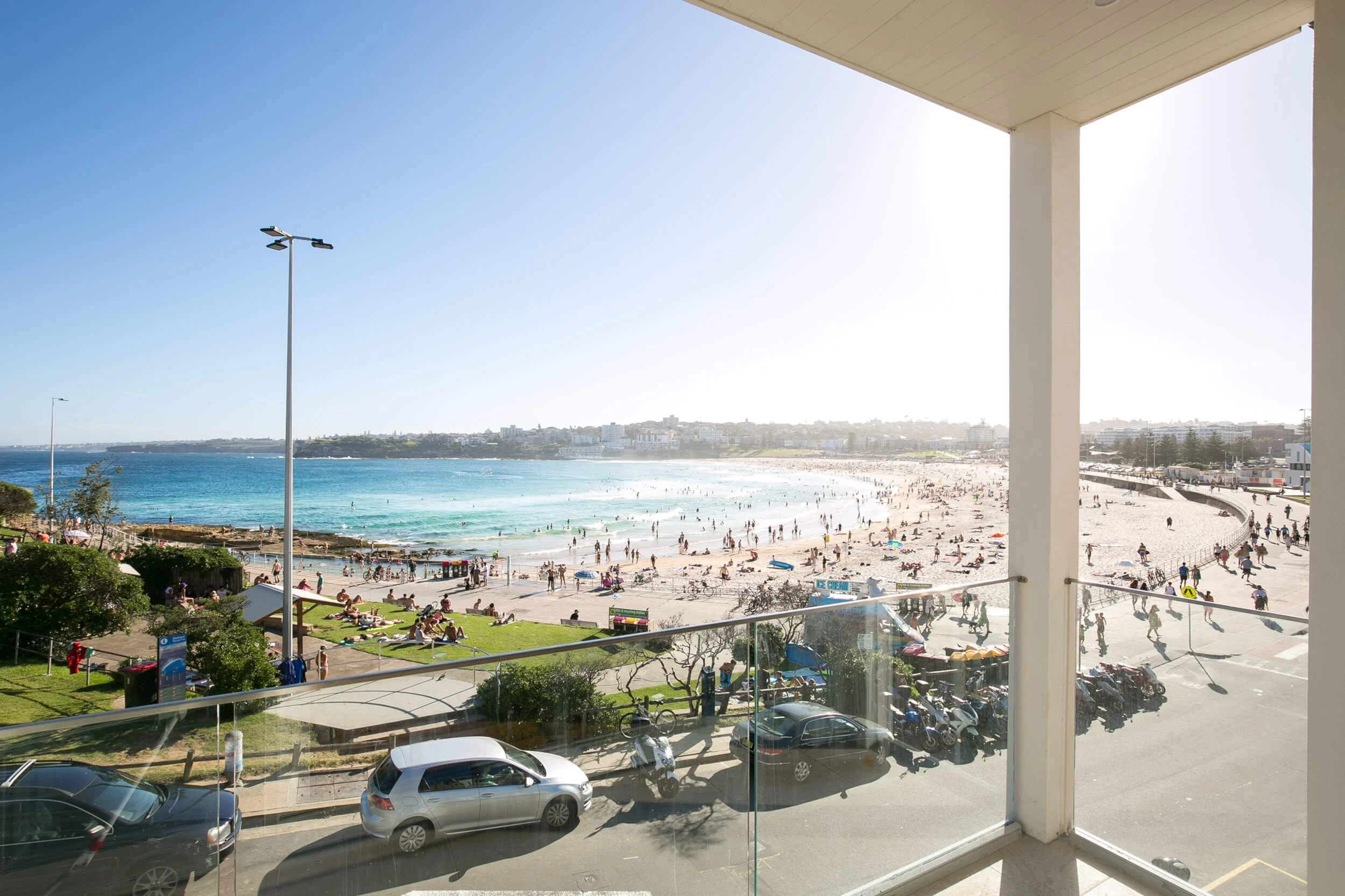 View of a busy beach from a balcony, with people sunbathing, swimming, and walking along the shoreline, and parked cars below.