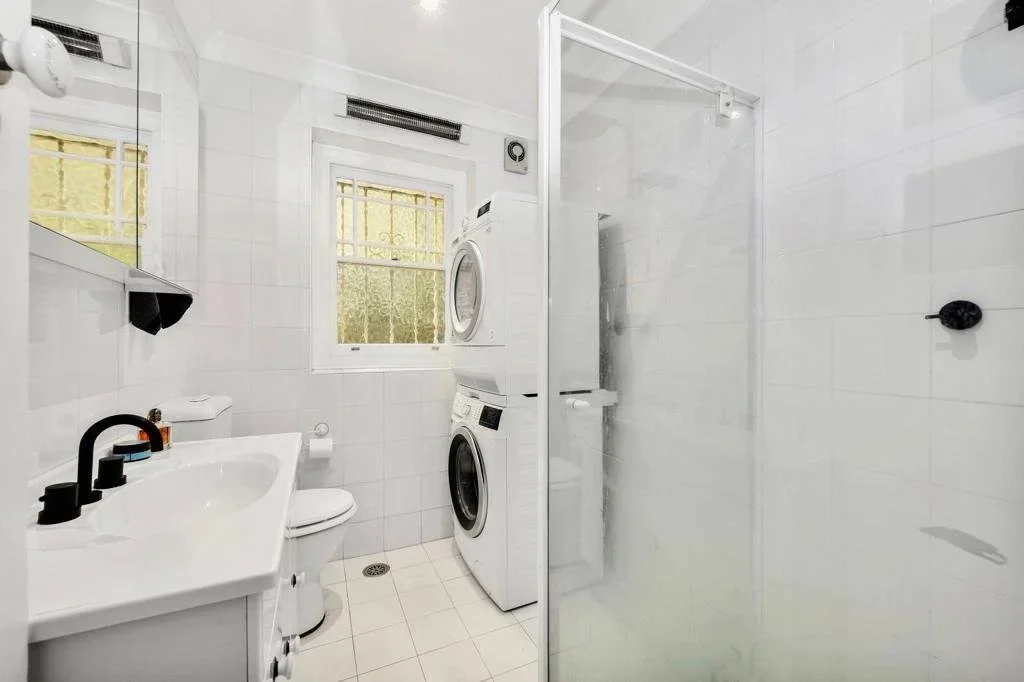 Small bathroom with white tiled walls and floor, featuring a sink with black fixtures, a toilet, two stacked washing machines, a frosted window, and a enclosed glass shower.