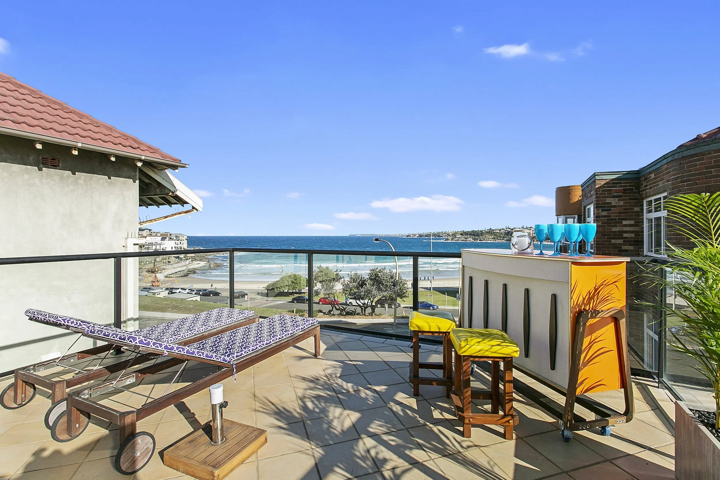 Balcony with a lounge chair, a bar with blue glasses, and view of the ocean, beach, and parking lot.
