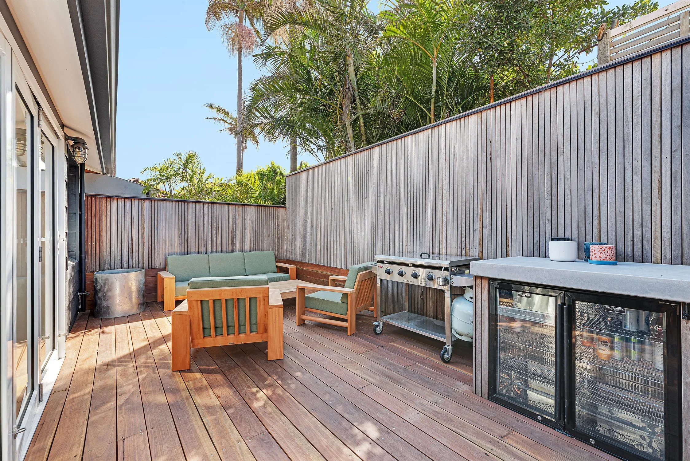 Outdoor patio with wooden flooring, green cushioned seating, a gas grill, a mini fridge with a transparent door, and tropical plants in the background.