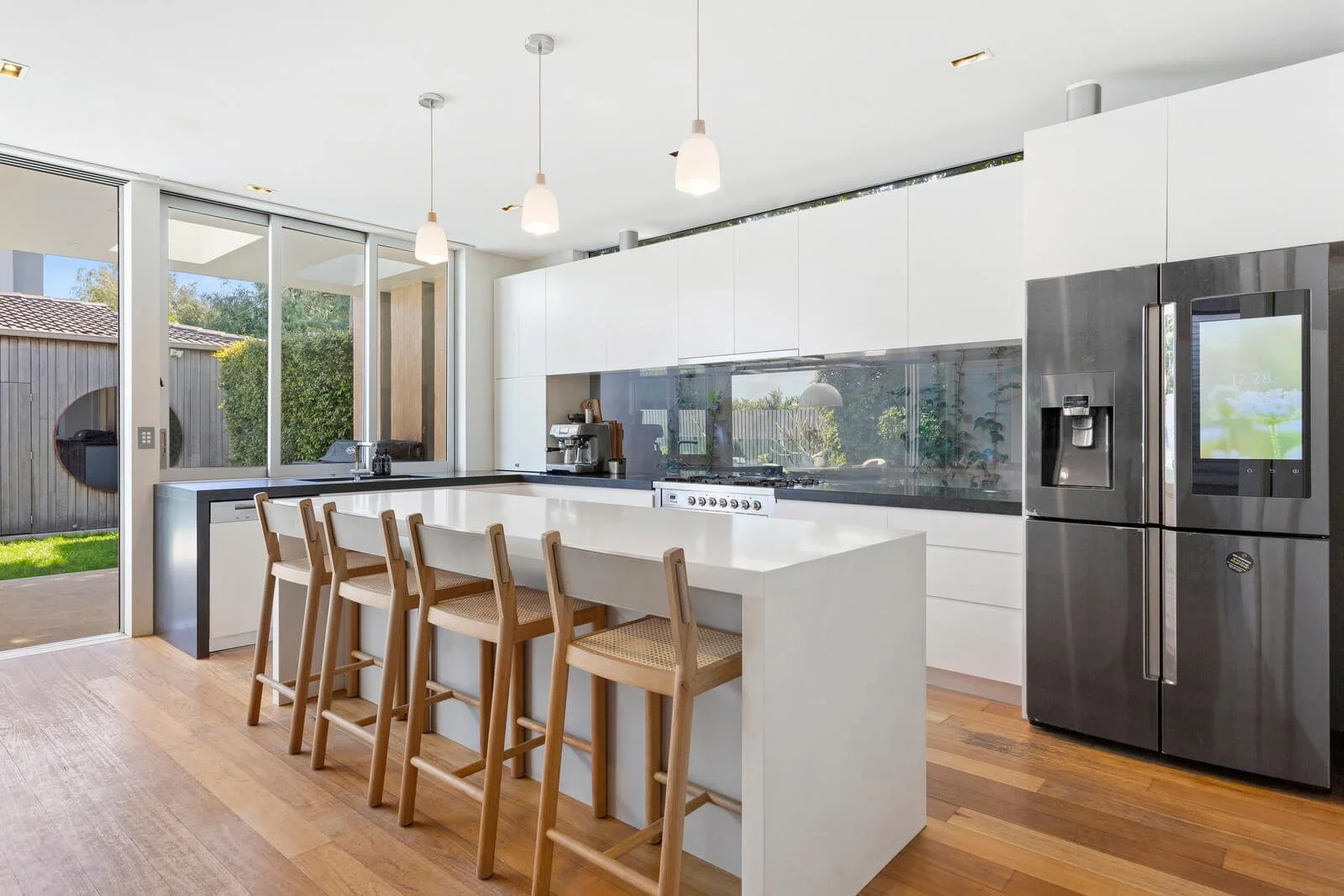 Modern kitchen with white cabinetry, black countertops, a stainless steel refrigerator, a kitchen island with four wooden chairs, hanging lights, and large windows showing a backyard with grass and a fence.