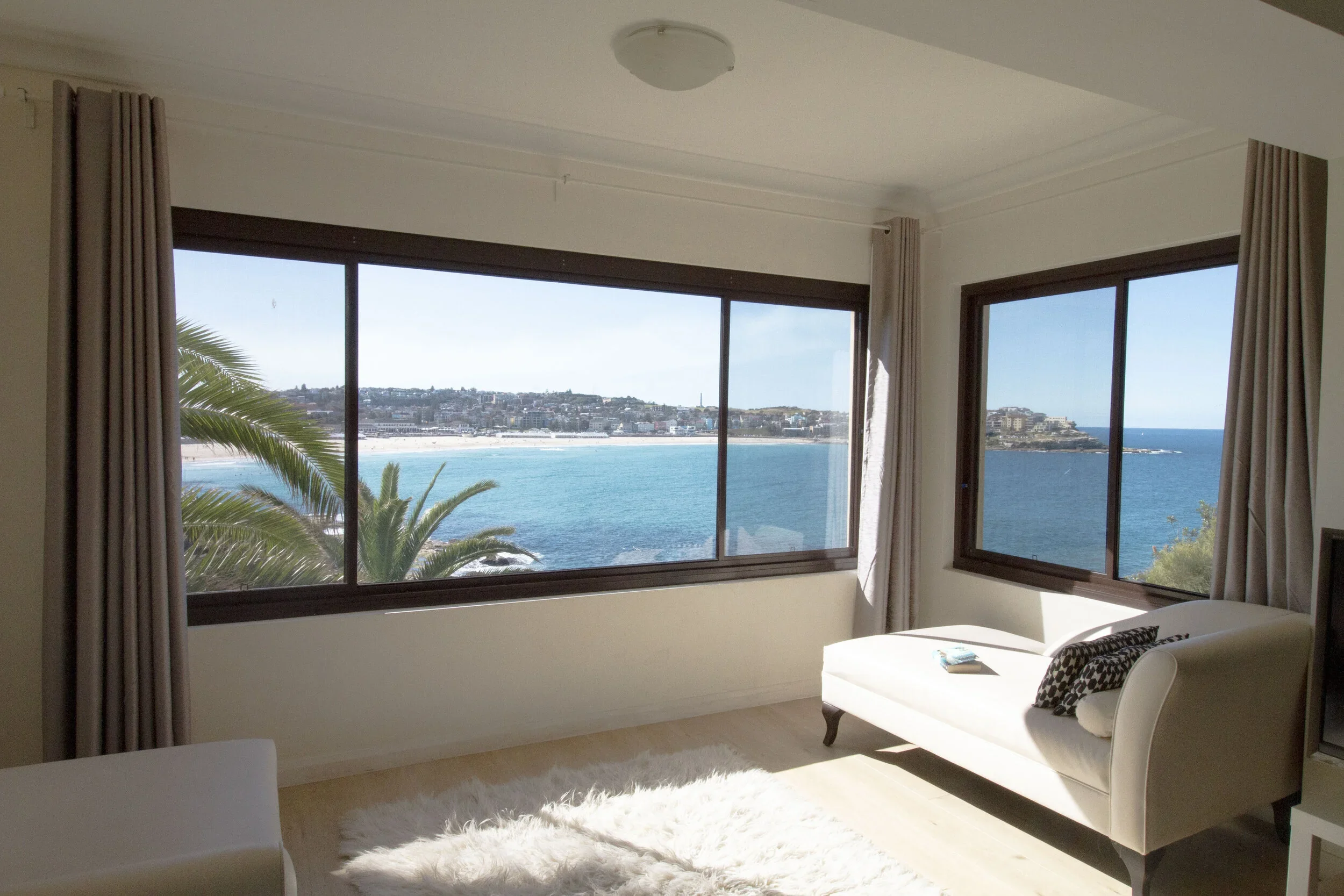 Living room with large windows overlooking the ocean and coastline, beige curtains, white sofa with patterned pillows, reading material on the sofa, and a fluffy white rug on a light wood floor.