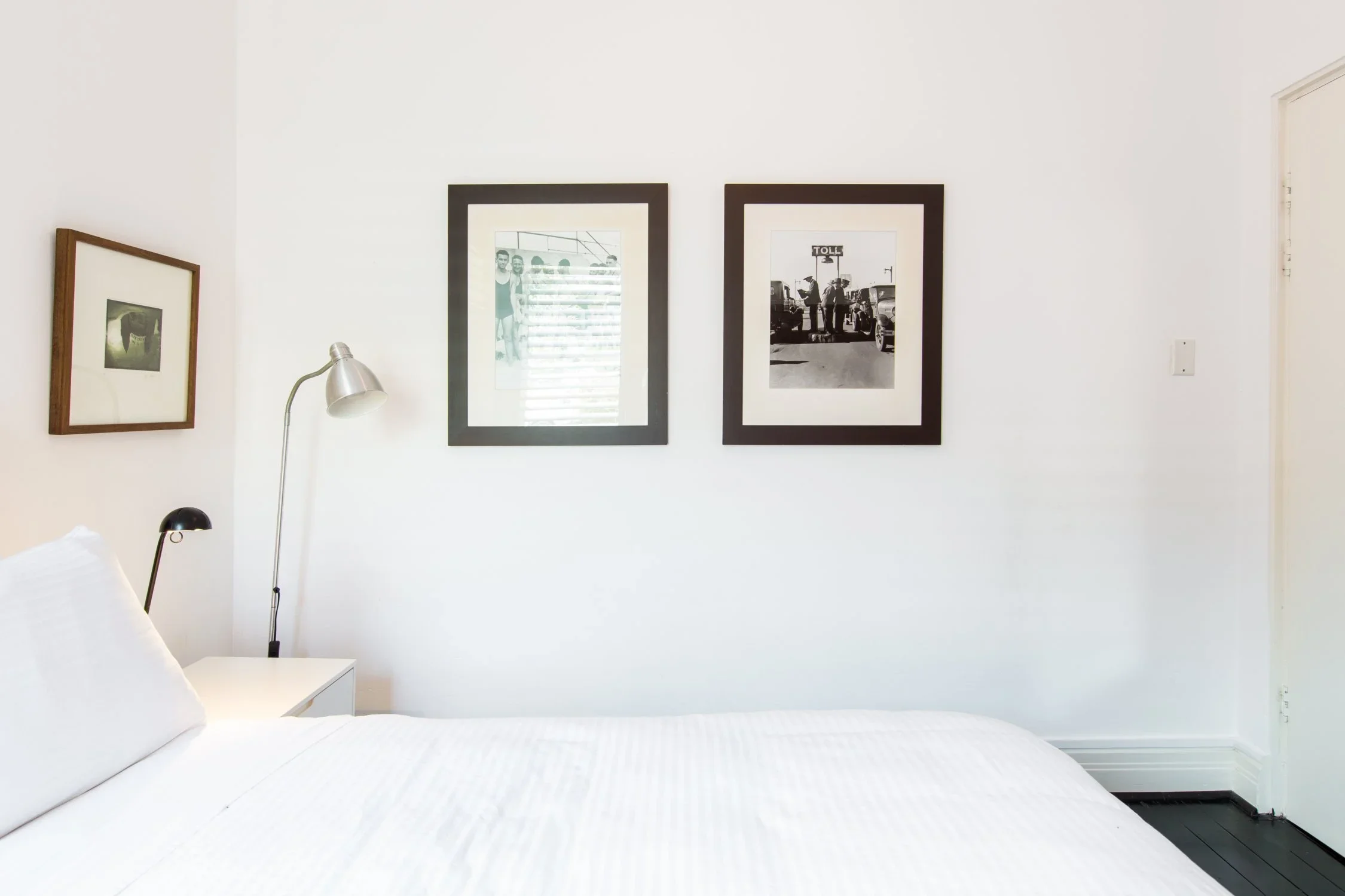 A bedroom with a white bed, black and silver wall lamps, three framed black and white photographs on a white wall, and a partially visible door on the right.