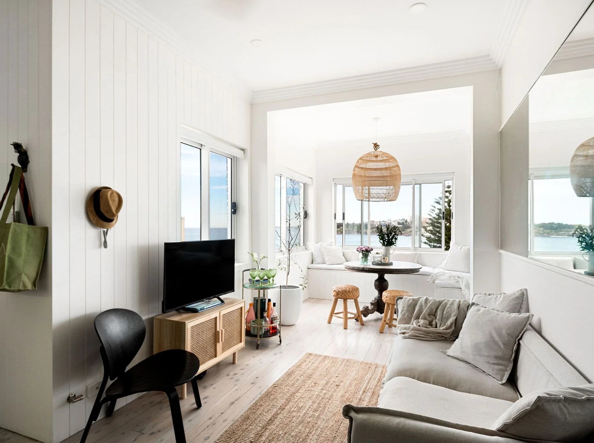 Bright living room with white walls, large windows showing a view of water and trees, a white sectional sofa with pillows, a round coffee table, a black chair, a flat-screen TV on a wooden stand, a hanging woven light fixture, some plants, and a wick