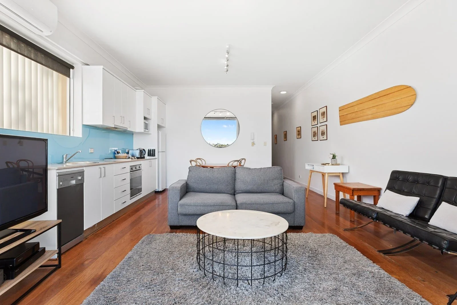 Living room with gray sofa, black leather lounge chair, round coffee table, TV, and an open kitchen area with white cabinets and blue backsplash.