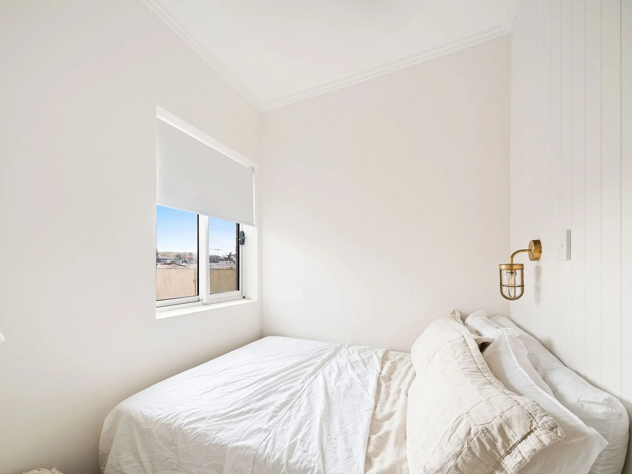 A minimalist bedroom with white walls, a window with a blind open to a city view, a bed with white bedding, and a wall-mounted brass and glass light fixture.