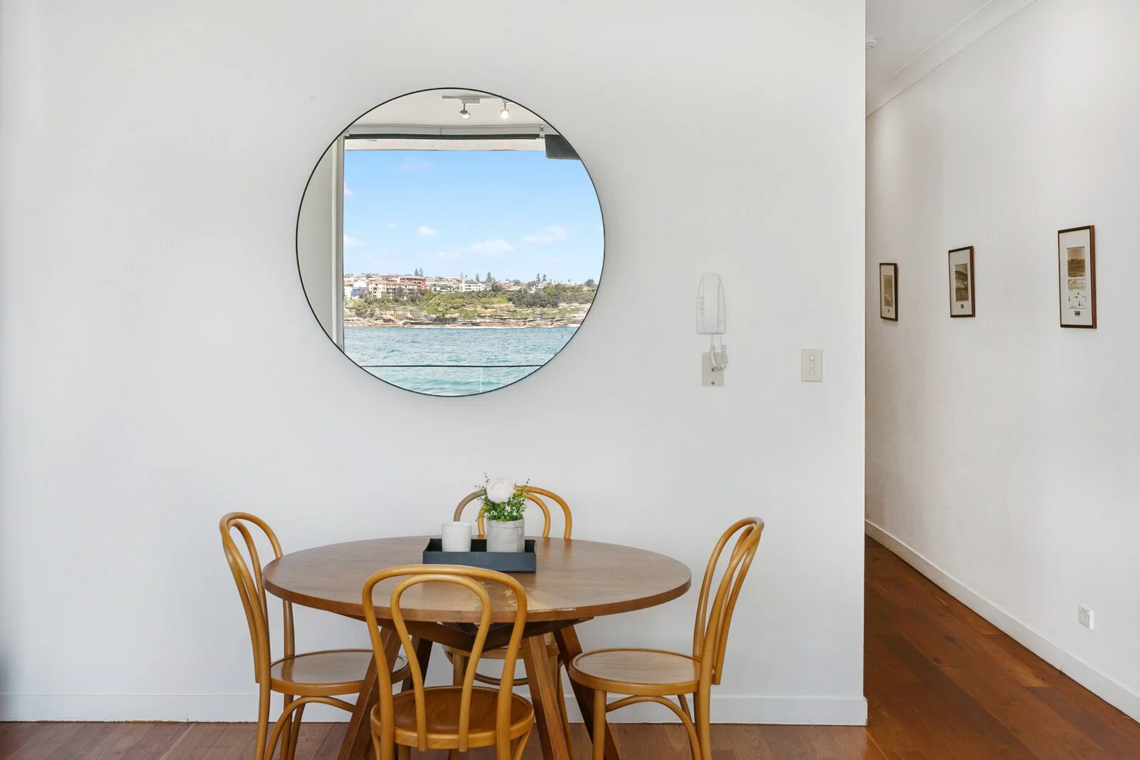 A dining area with a round wooden table and four matching chairs. On the table, there is a tray with a flower vase and candles. A large, round mirror on the white wall reflects a window with a view of the ocean and buildings on a hill. There are thre