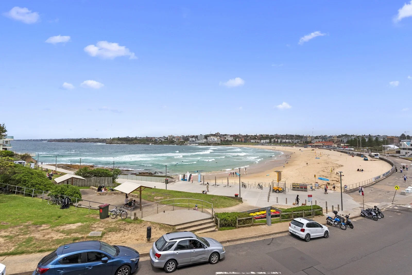 A beach scene with a sandy shore, ocean waves, a promenade, parked cars, bicycles, and people enjoying the weather under a blue sky with scattered clouds.