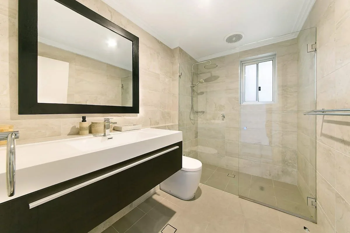 Modern bathroom with a large mirror, white sink vanity with black drawer, white toilet, shower area with glass enclosure, rainfall showerhead, small window, and beige tiled walls and floor.