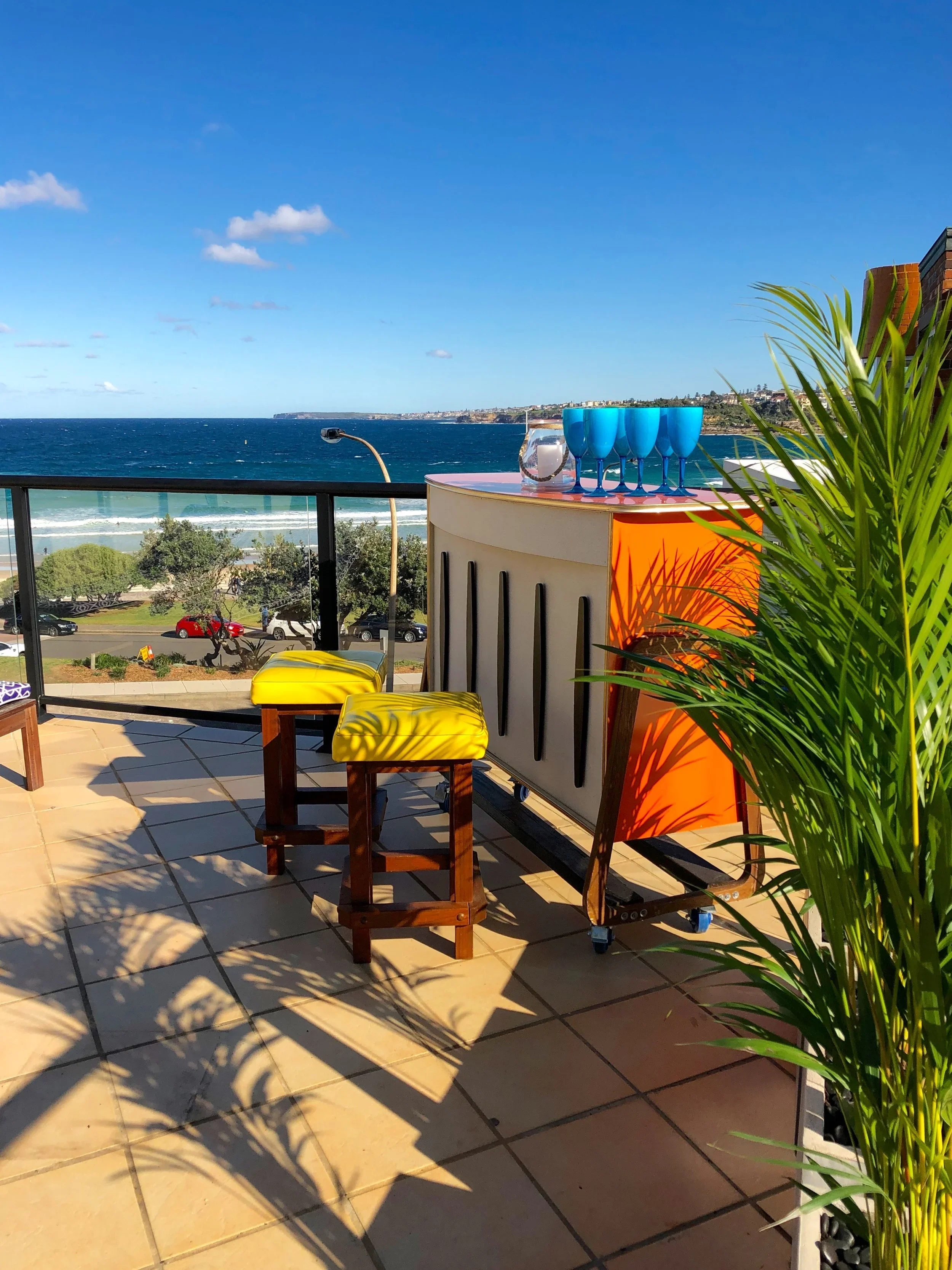 Balcony with a view of the ocean, featuring a bar with four blue glasses, a water pitcher, two yellow cushioned stools, and a green potted plant, under a sunny sky.