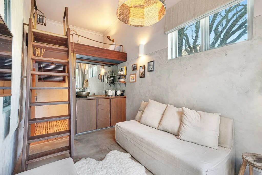 Small living space with a white sofa, wooden loft bed accessed by a ladder, compact kitchen with open shelves, wall art, a large window with blinds, and a ceiling light fixture.
