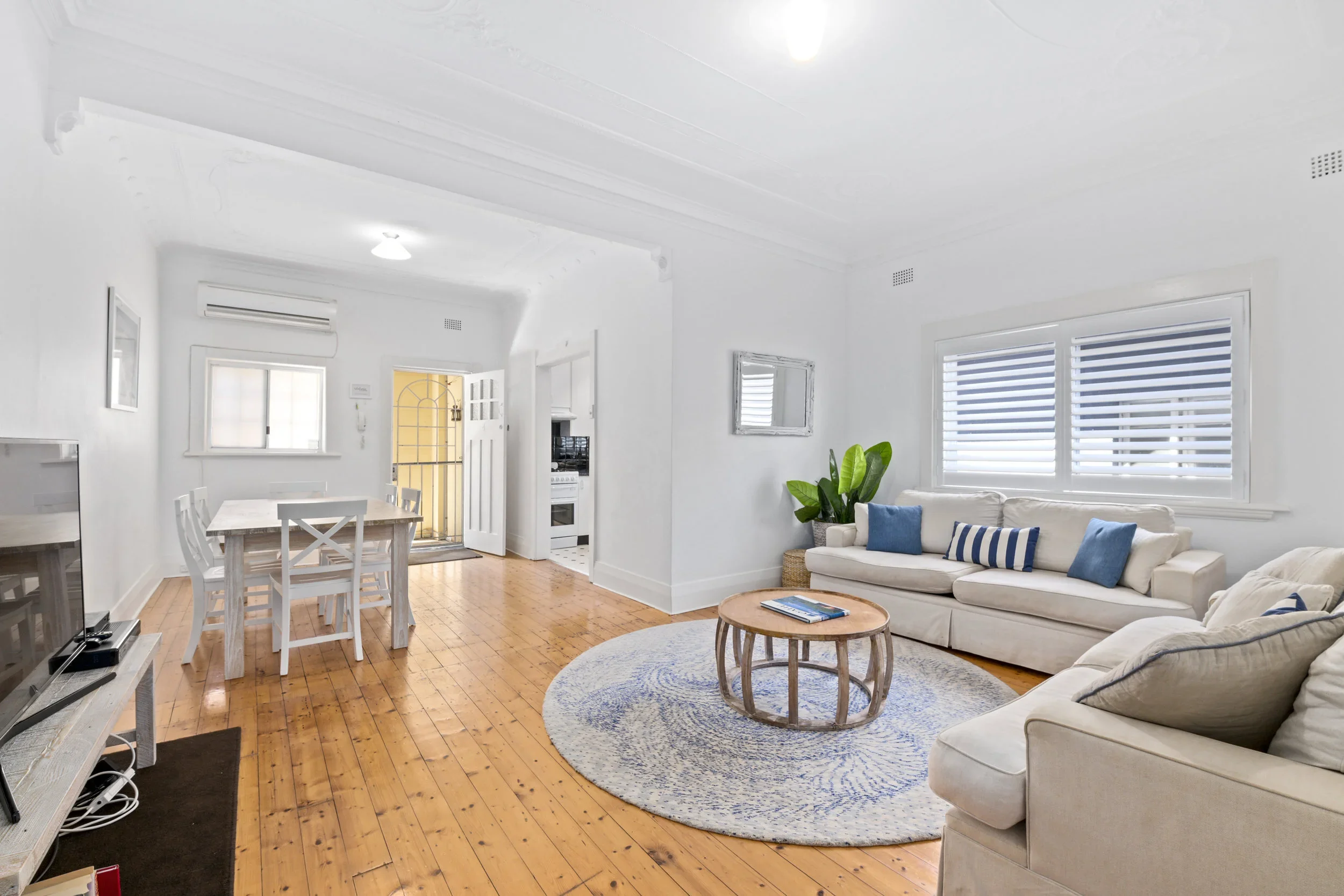 Bright living room with white walls, wooden floor, beige sofas with blue pillows, a round wooden coffee table on a patterned rug, large window with shutters, and a dining area with a white table and chairs.