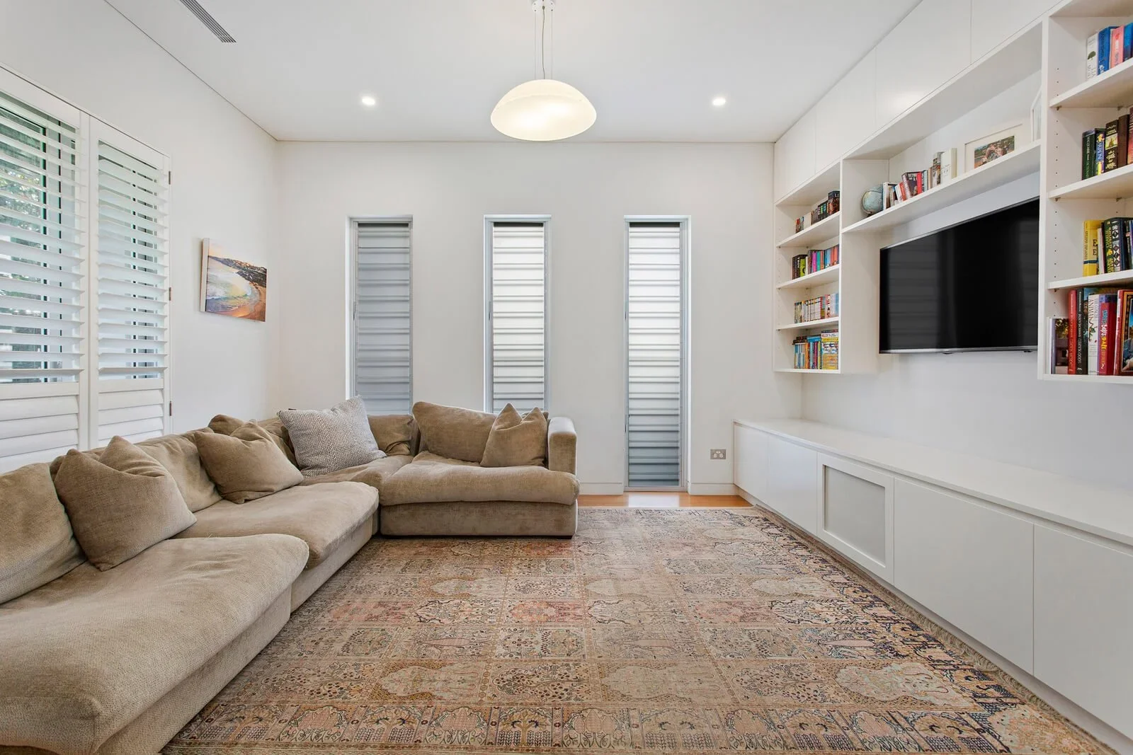 A contemporary living room with a beige sectional sofa, colorful patterned rug, white built-in shelf with books, and three narrow windows with blinds.