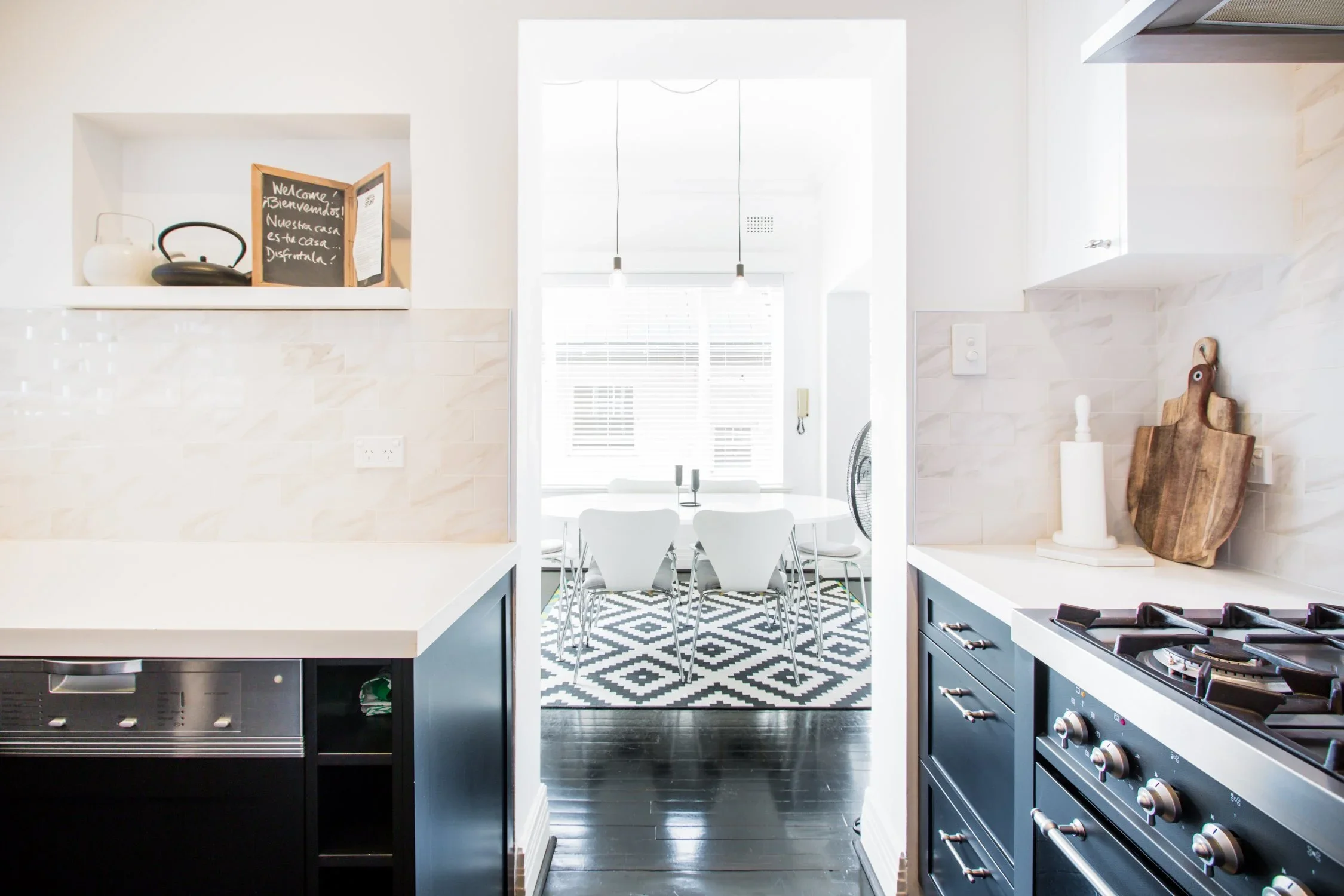 View of a modern kitchen opening to a bright dining room with white chairs and a patterned black-and-white rug.