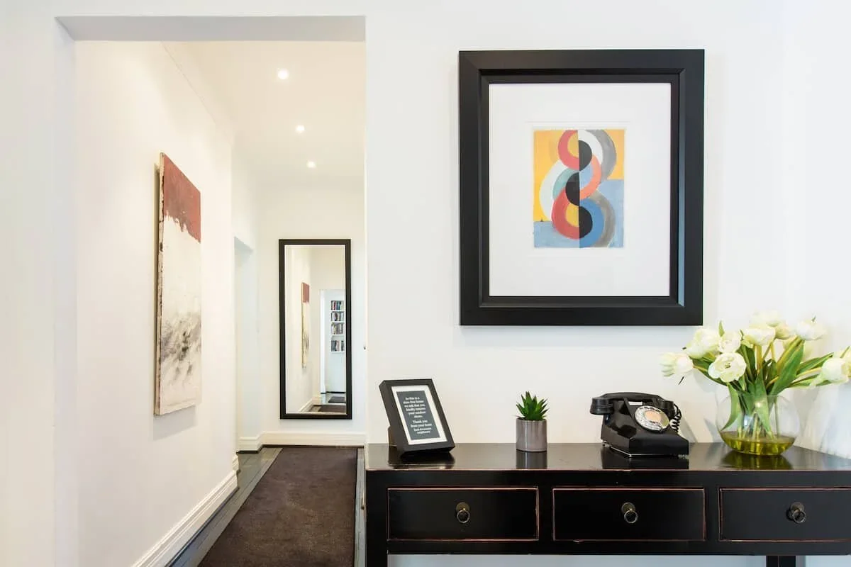 Modern hallway with white walls, black furniture, colorful abstract art, a mirror, a vintage rotary phone, and a vase with white flowers.