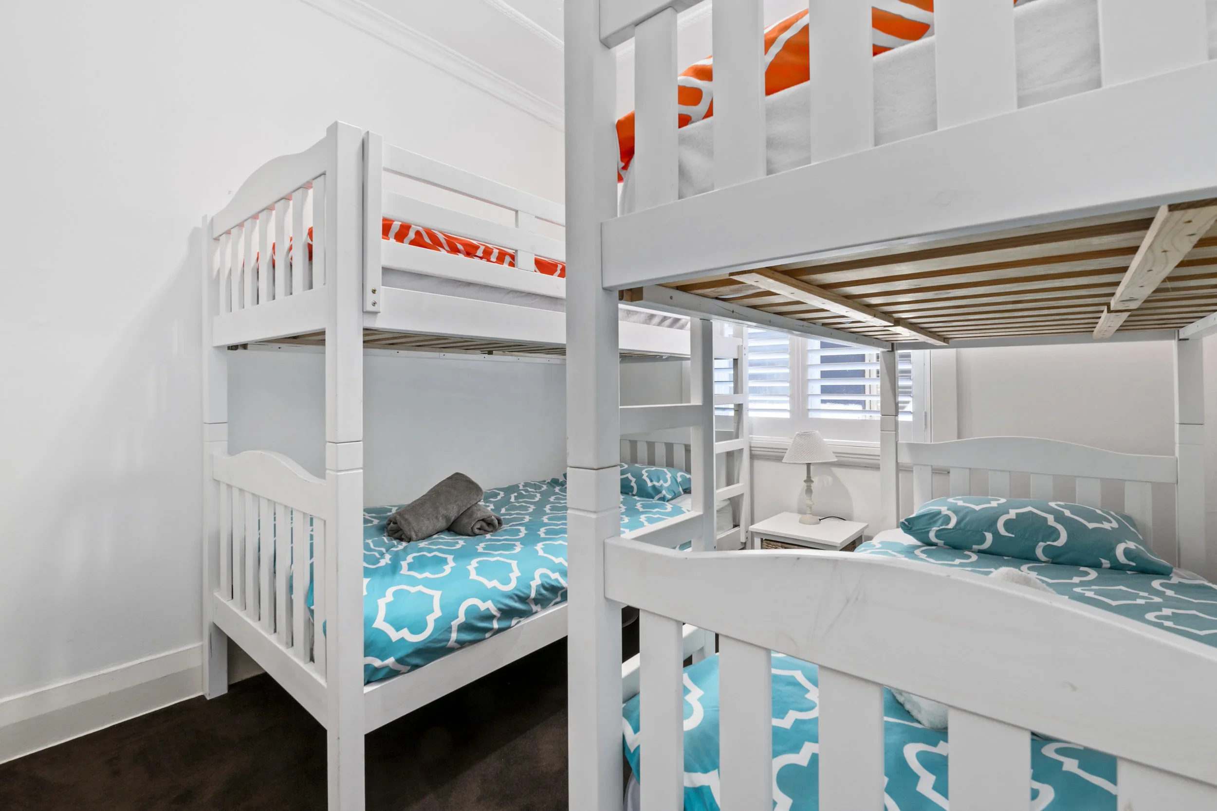 A bedroom with two white bunk beds, blue patterned bedding, a nightstand with a lamp, and windows with white blinds.
