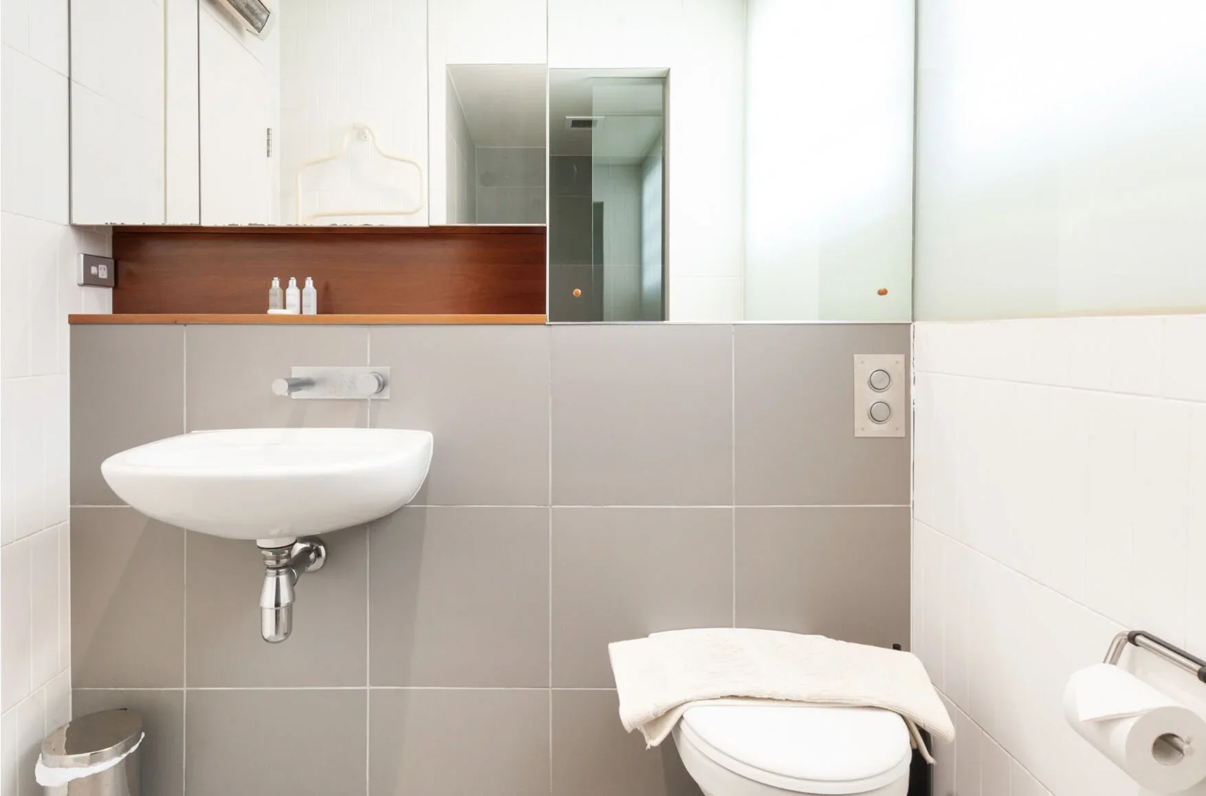 Small bathroom with a white sink, toilet with a towel on top, and a mirror above the sink. There are bottles of toiletries on a wooden shelf and a frosted glass window letting in natural light.