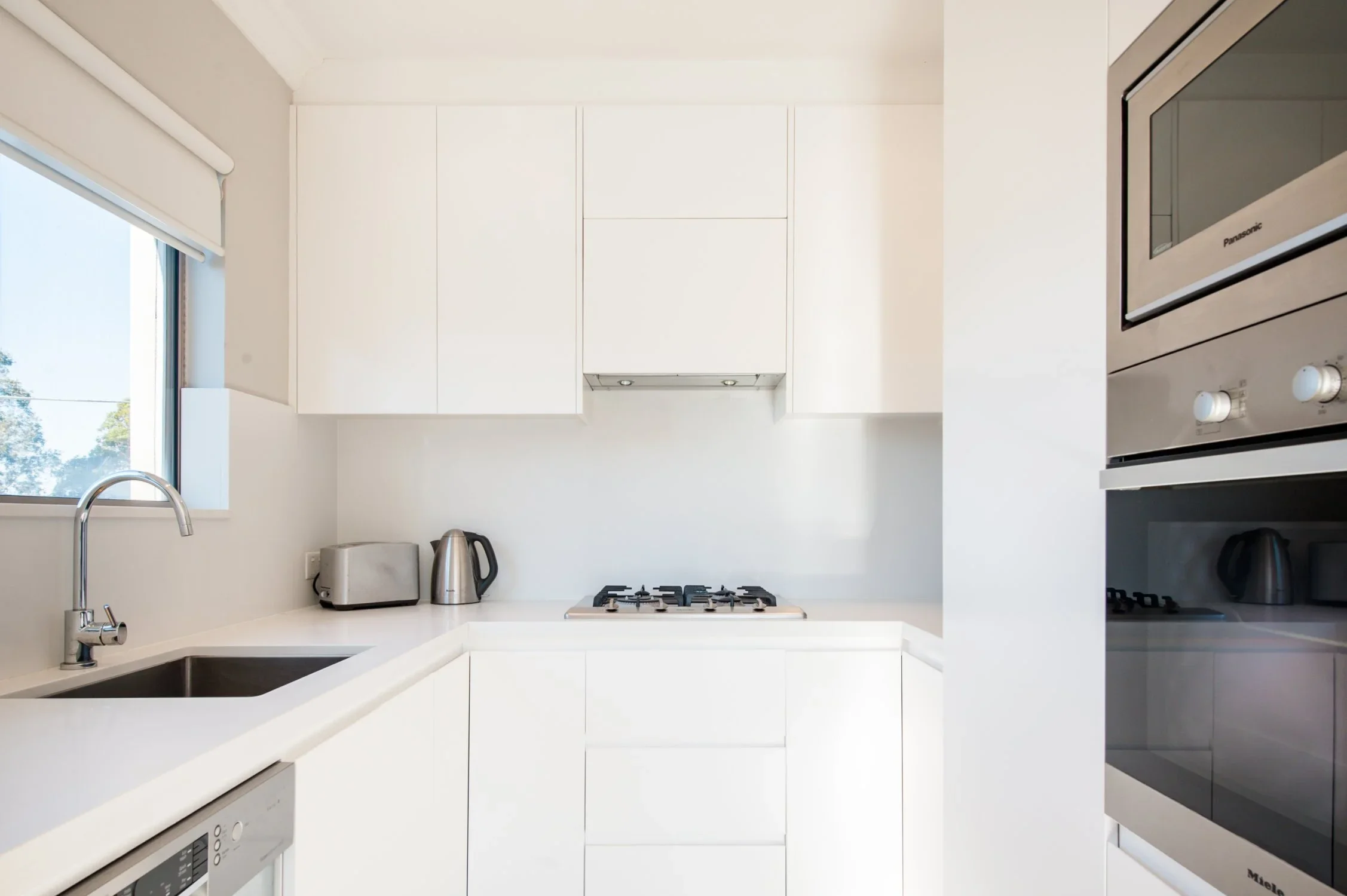 Minimalist white kitchen with window, sink, modern appliances, and cabinets.