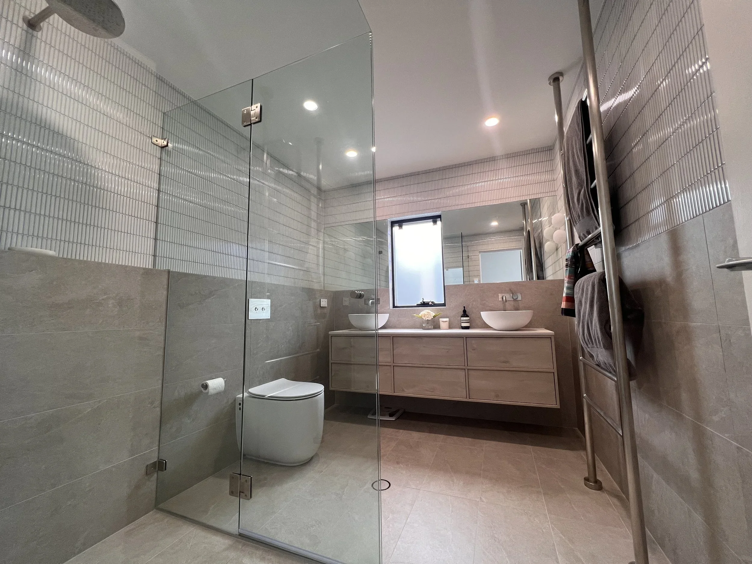 Modern bathroom with a glass-enclosed shower, a wall-mounted toilet, and a vanity with two vessel sinks. There is a window, a large mirror, and a heated towel rail with towels.