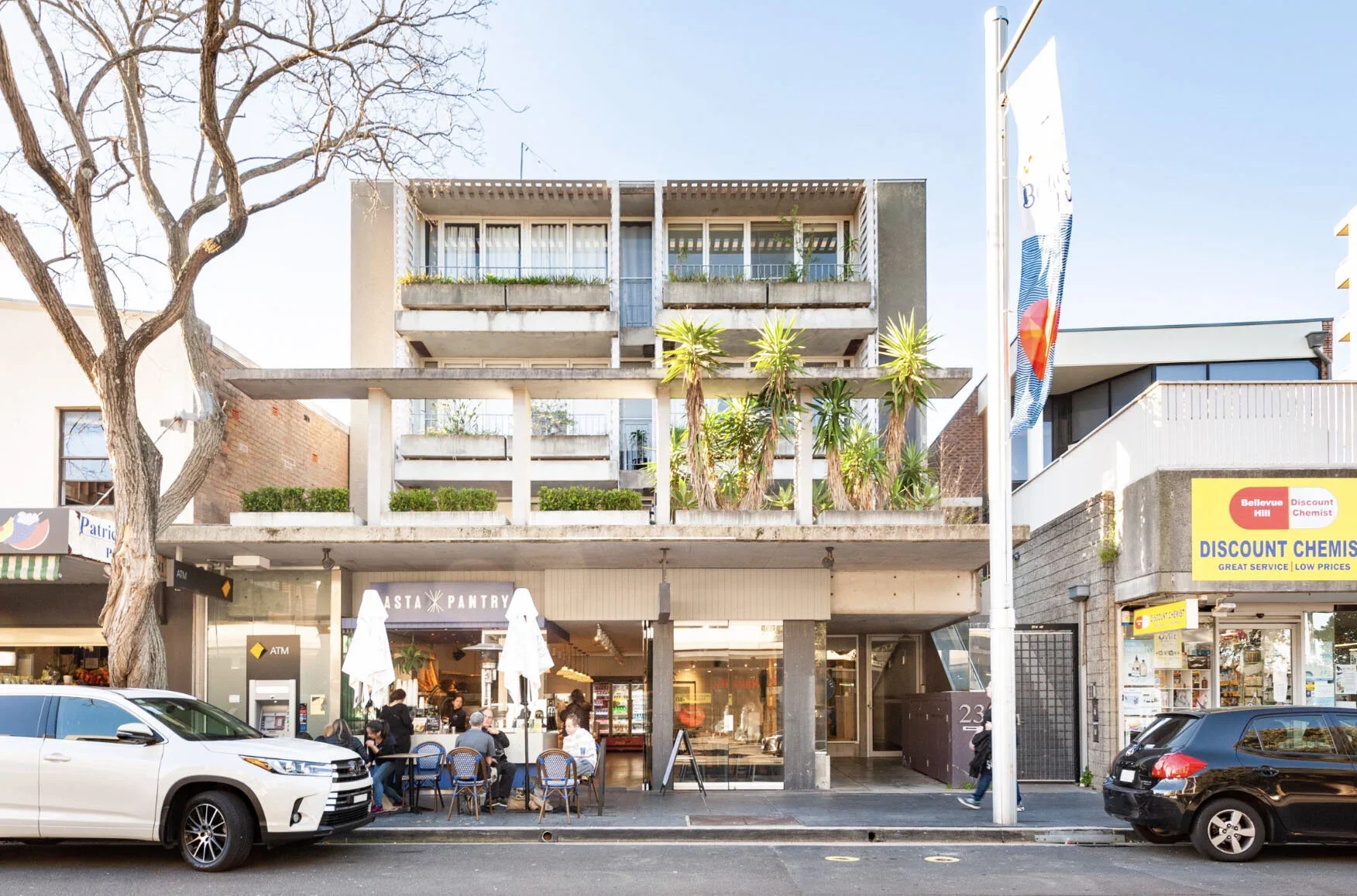 A multi-story building with a bakery or cafe on the ground floor and residential or office space above, with outdoor seating and greenery on the second level, parked cars, and pedestrians on the street.
