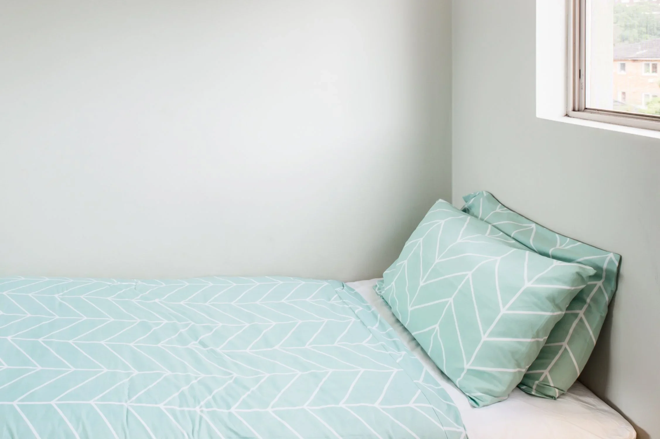 A bedroom with a bed featuring light blue and white leaf-patterned sheets and pillowcases, positioned near a window with view of buildings outside.
