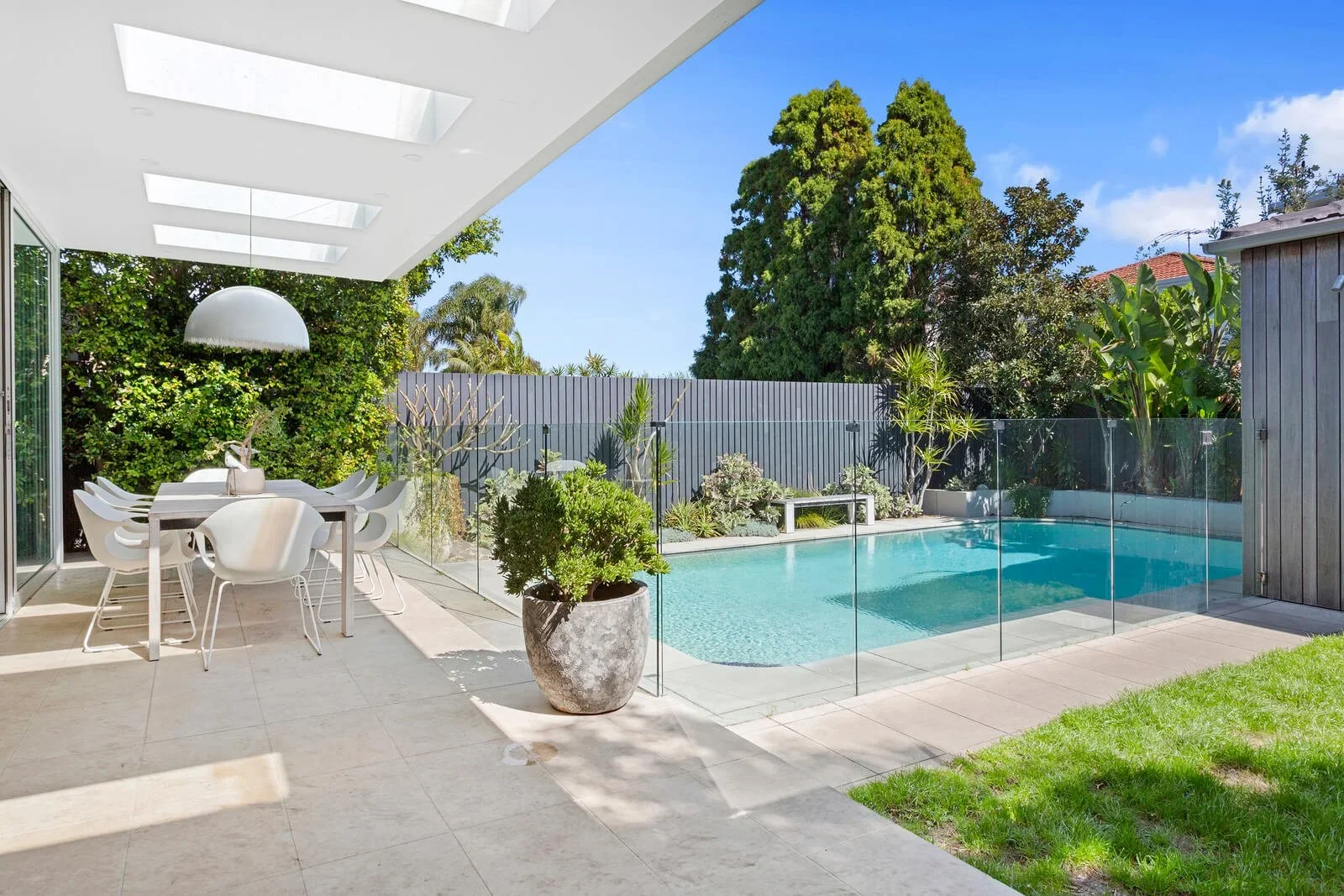 Modern backyard with a swimming pool, outdoor dining area with white chairs and table, potted plant, and surrounded by greenery and trees.