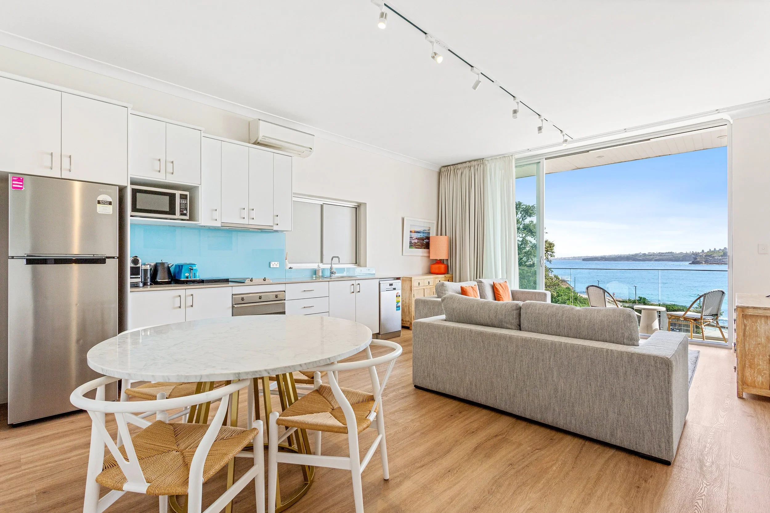 Open-concept living room and kitchen with view of ocean through large sliding glass door, light-colored sofas, dining table with four chairs, and kitchen appliances including refrigerator, microwave, and coffee maker.