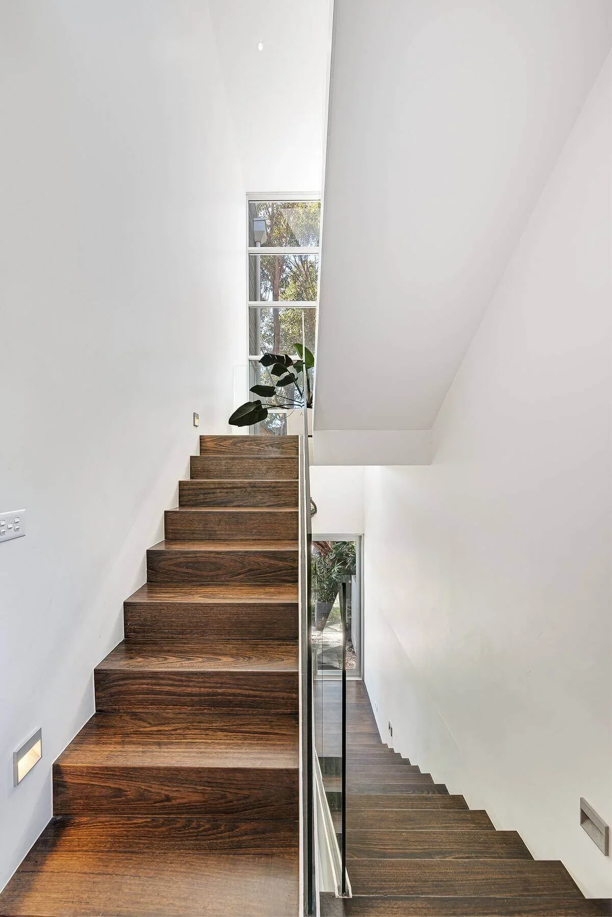 Modern staircase with dark wood steps, glass railing, white walls, and large window at the top providing natural light.