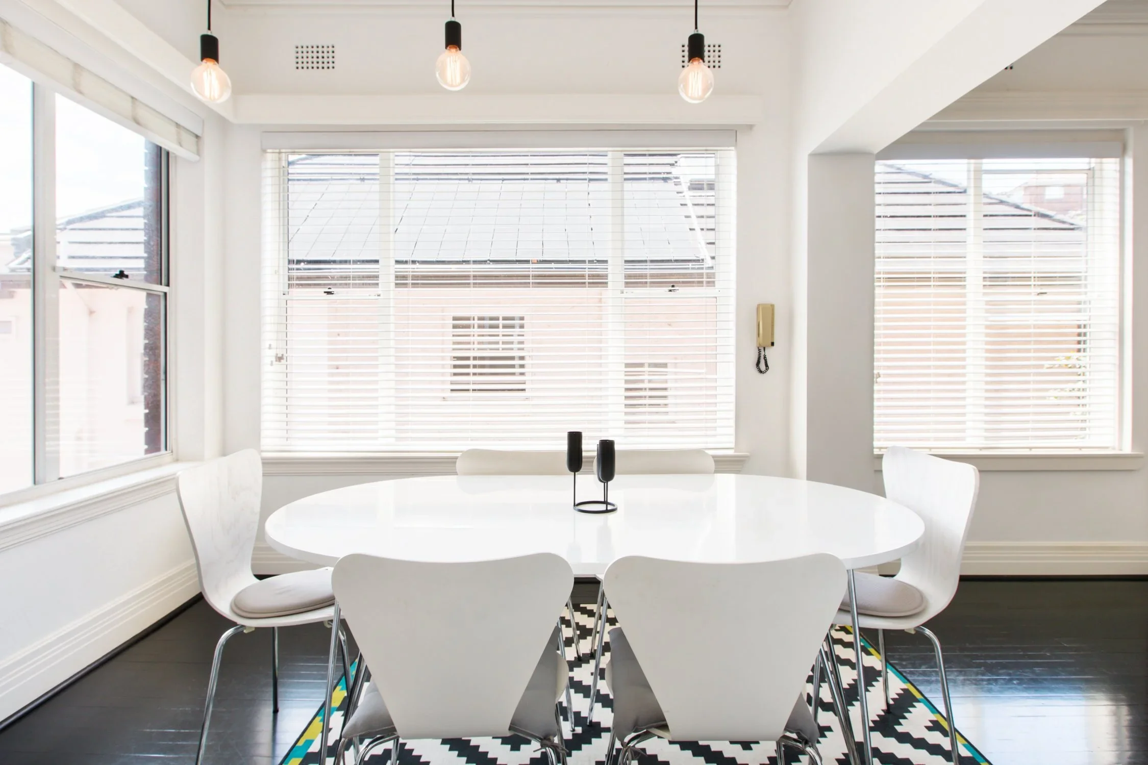 Bright dining room with large windows covered with blinds, white walls, and a white oval table with six white chairs. The room has black flooring, a geometric black and white rug, hangers with bulbs and a wall-mounted telephone.