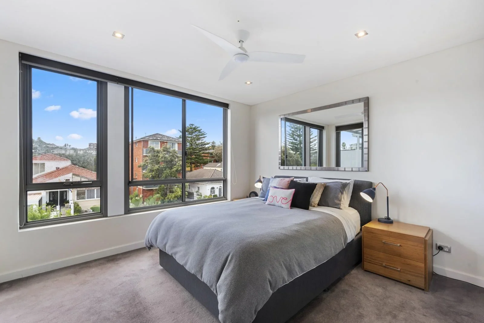 A bedroom with a large window showing a view of houses and trees, a bed with pillows and a gray blanket, a mirror on the wall, a wooden nightstand with a small lamp, a ceiling fan, and recessed ceiling lights.