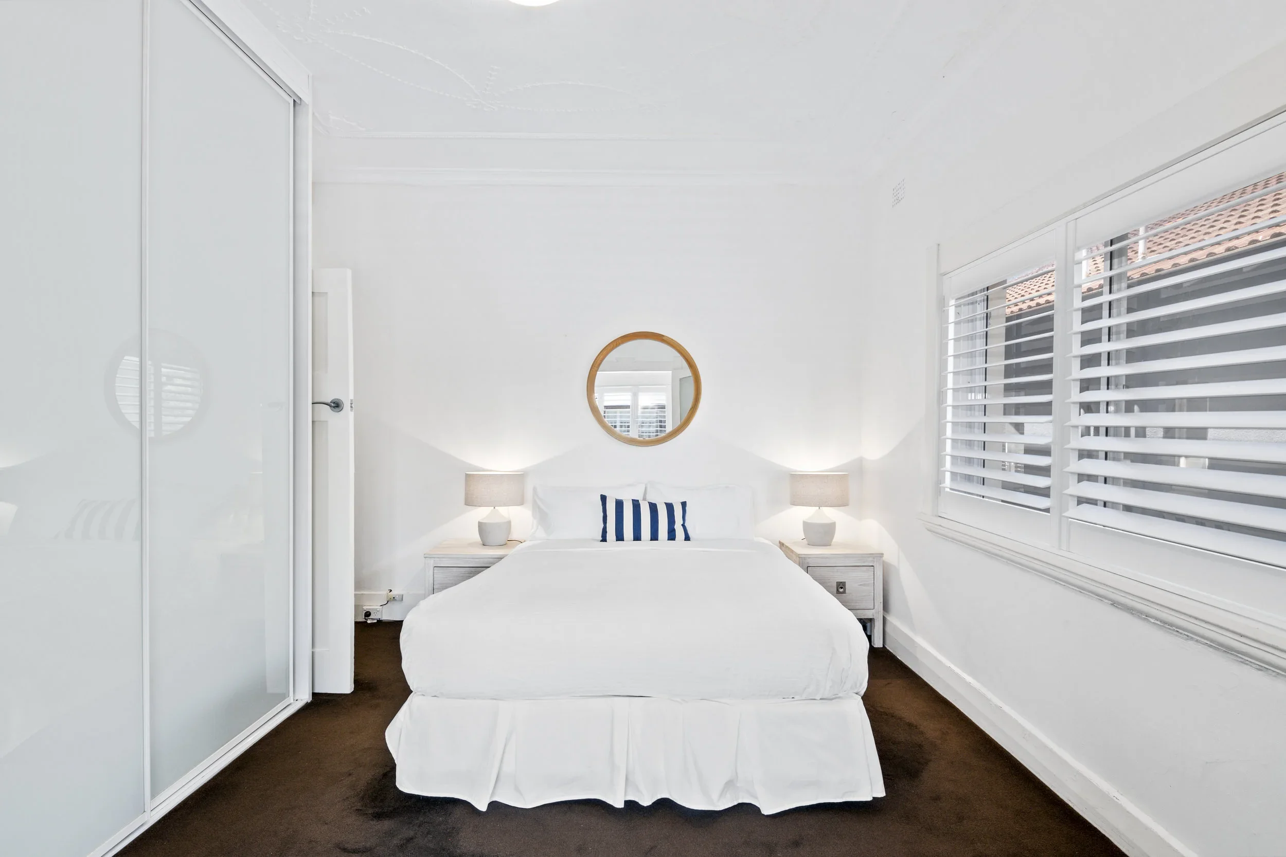 A bright, white bedroom featuring a neatly made bed with a blue striped pillow, flanked by two nightstands with white lamps. A circular mirror hangs above the bed, and there are large windows with white blinds on the right.