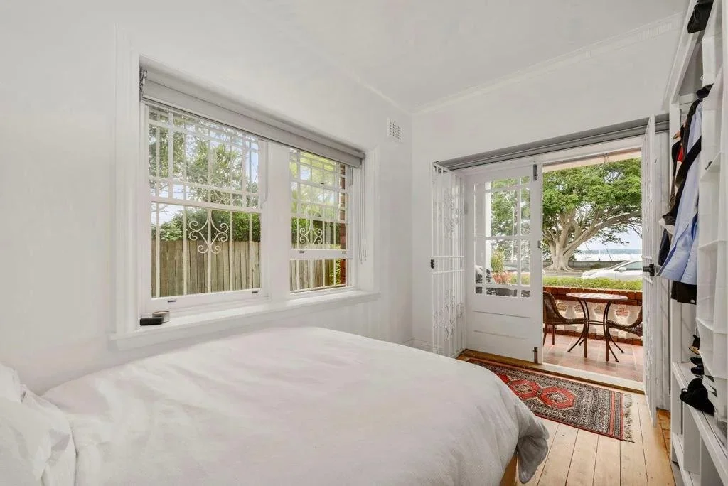 Bedroom with white walls, a bed, a window with a decorative metal grill, and a door leading outside to a porch with a table and chairs near a waterfront view with trees.