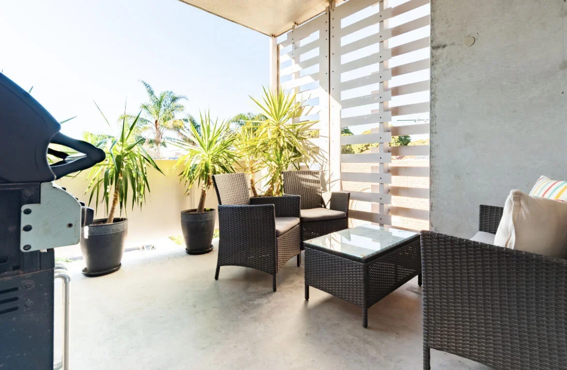 Balcony with black wicker chairs, a glass-topped table, large potted tropical plants, and a white wood privacy screen, overlooking palm trees in bright sunlight.