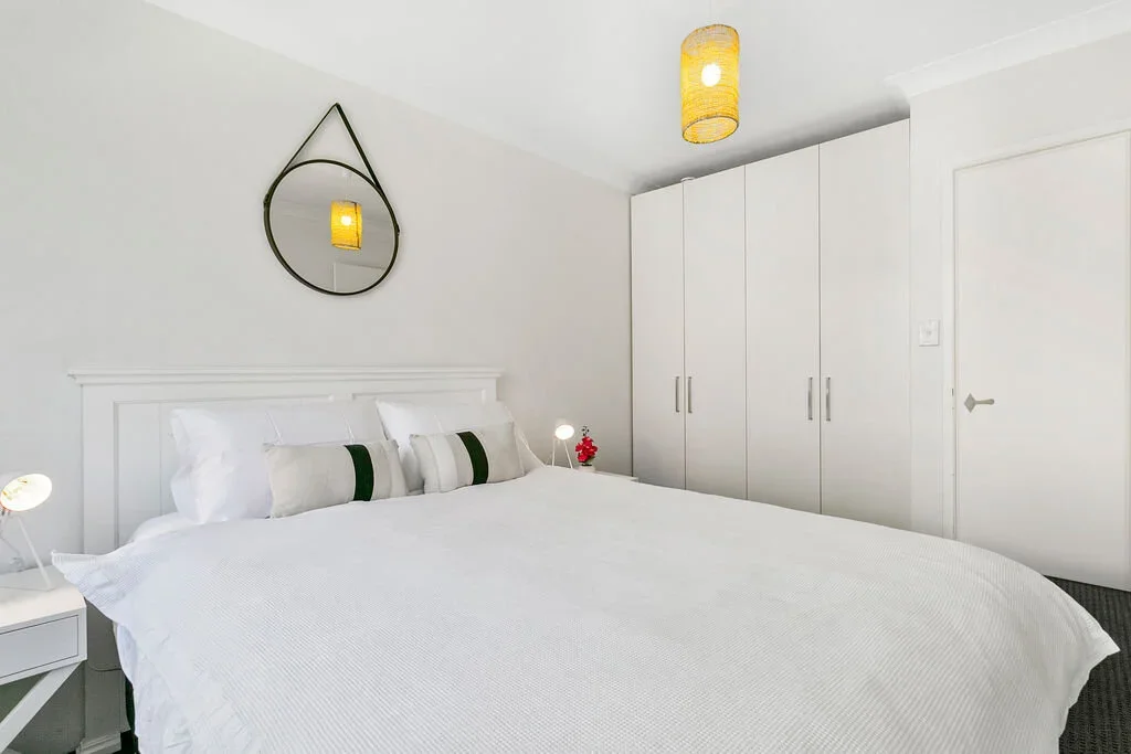 A minimalist bedroom with white walls, a large bed with white bedding and decorative pillows, a round mirror with a black frame and hanging strap, a white nightstand with a small lamp, a red flower, and a modern ceiling light fixture. There is a whit