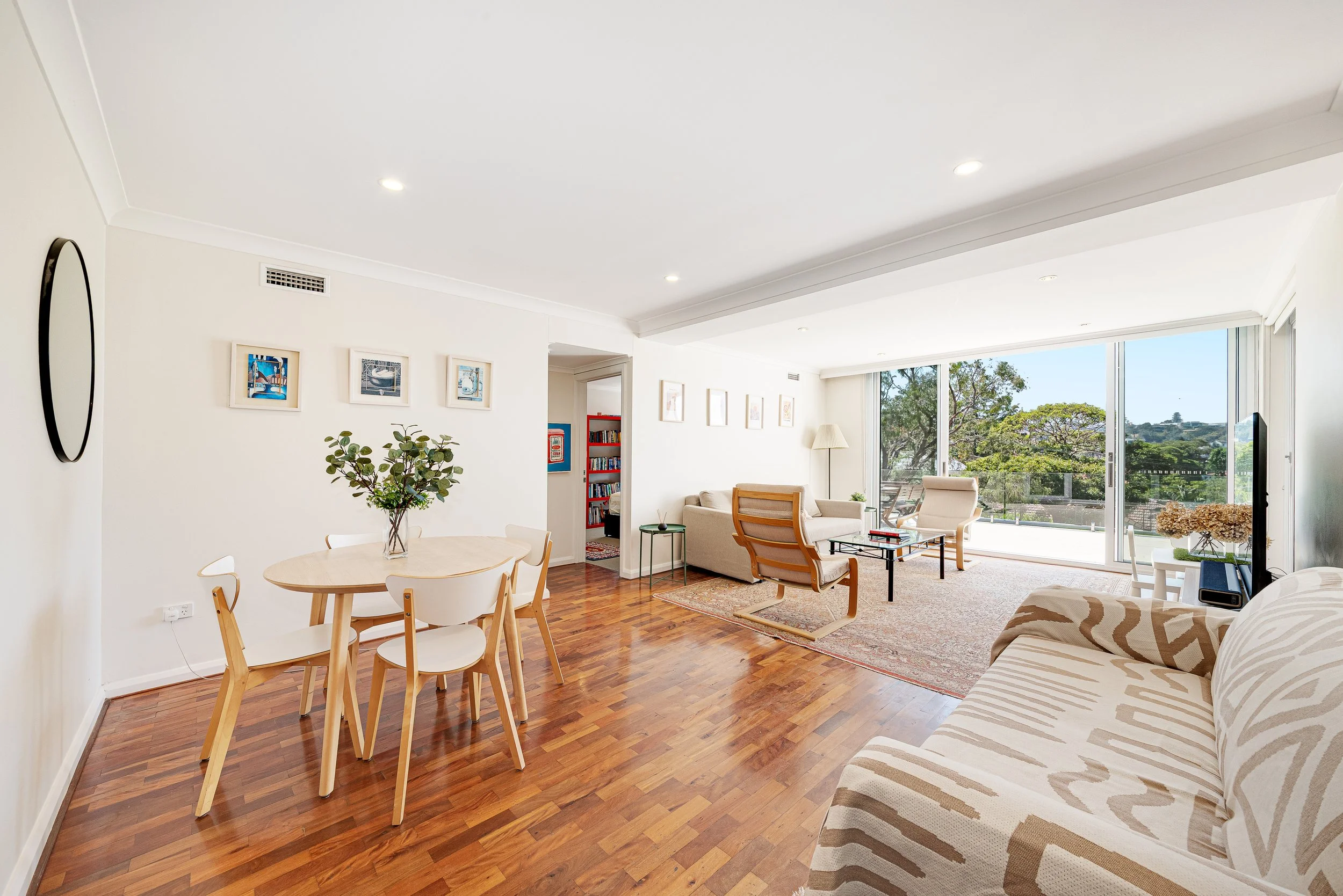 Bright living room with light-colored sofas, wooden flooring, a large sliding glass door leading to a balcony with outdoor seating, a television, and minimalist decor including framed pictures on the walls.