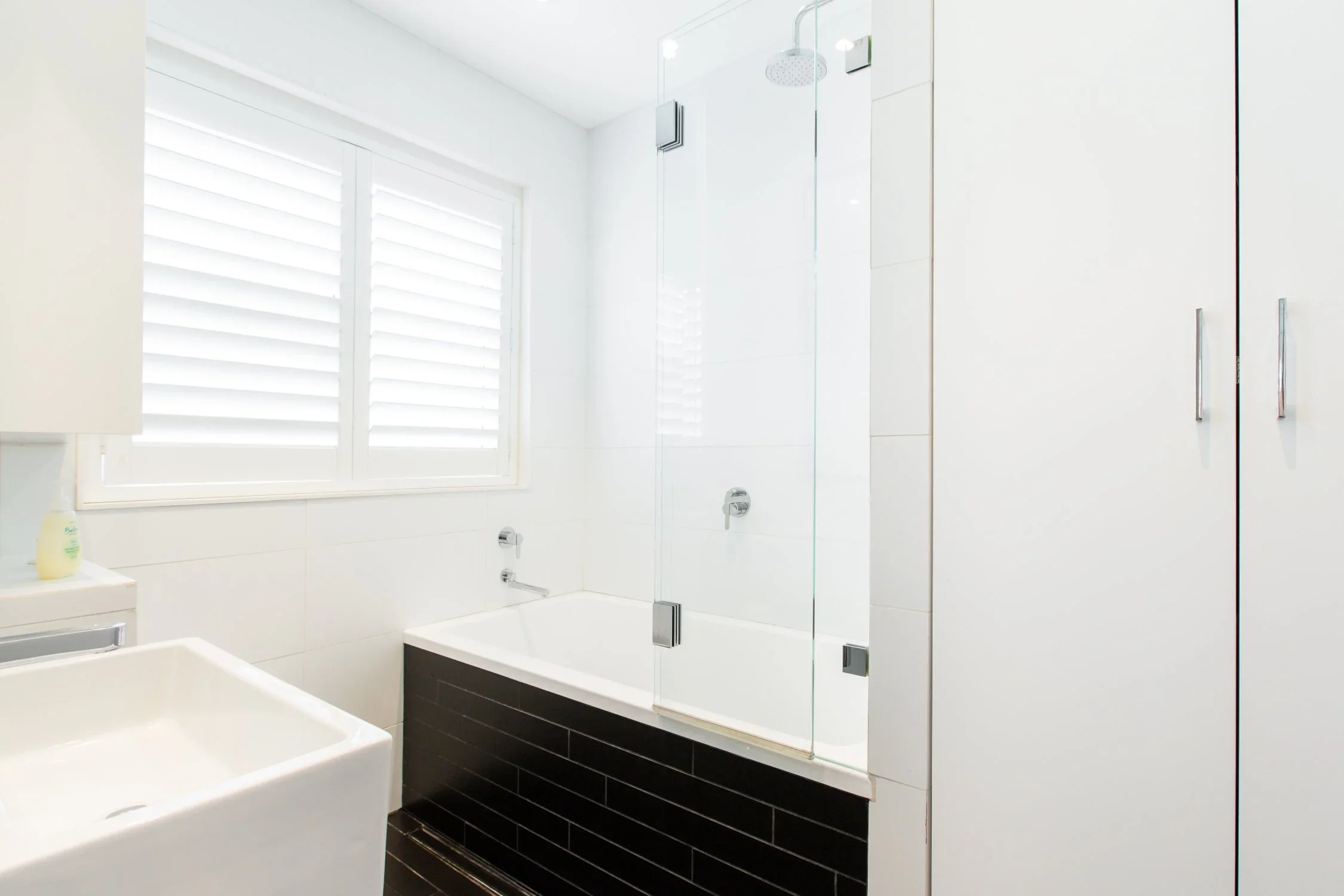 Modern bathroom with white walls, a window with horizontal blinds, a white bathtub with black tile surround, a glass shower enclosure, and white storage cabinets.
