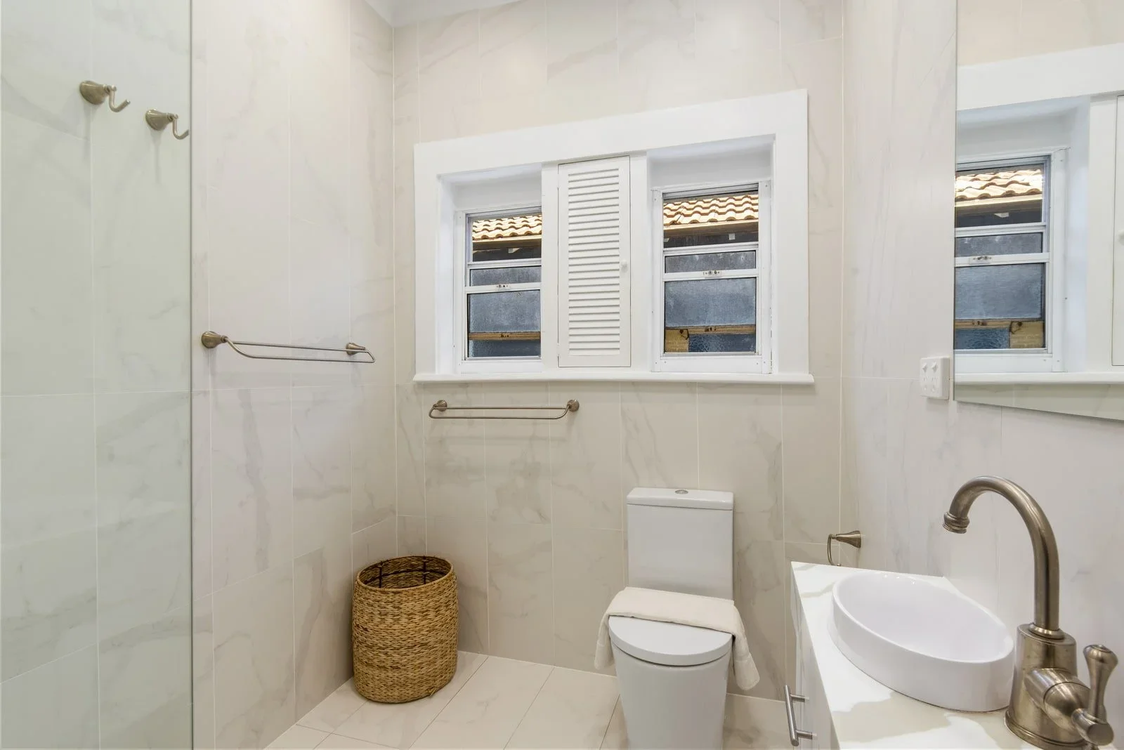 Clean bathroom with beige tiled walls, a small window, a toilet, a woven laundry basket, and a white sink with a brushed nickel faucet.